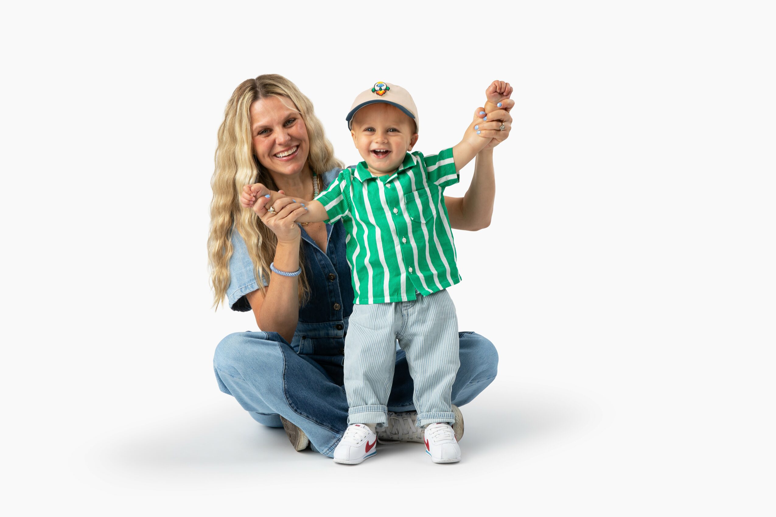 Woman and child sitting on the floor, smiling, holding hands, child wearing a striped green shirt and cap, white background.