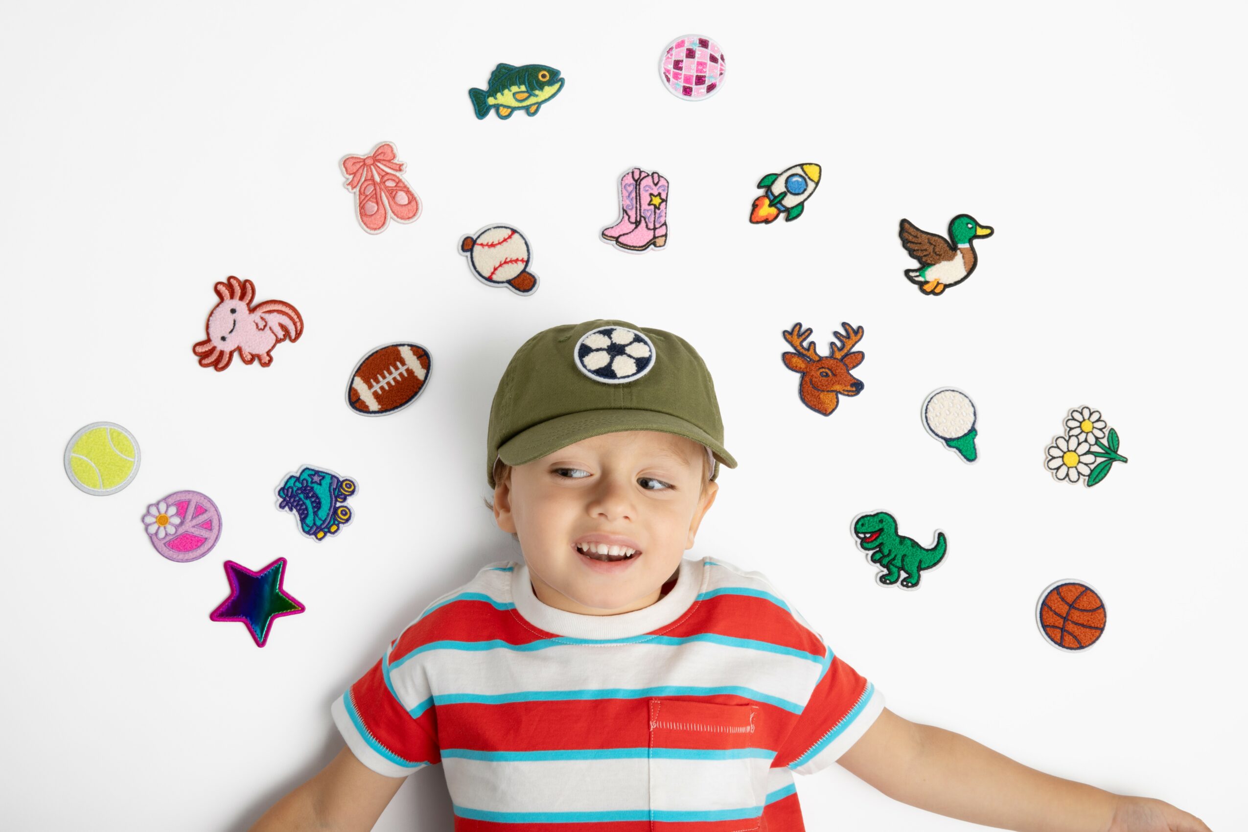 Child lying on white background surrounded by colorful stickers, wearing a green cap and striped shirt, smiling.