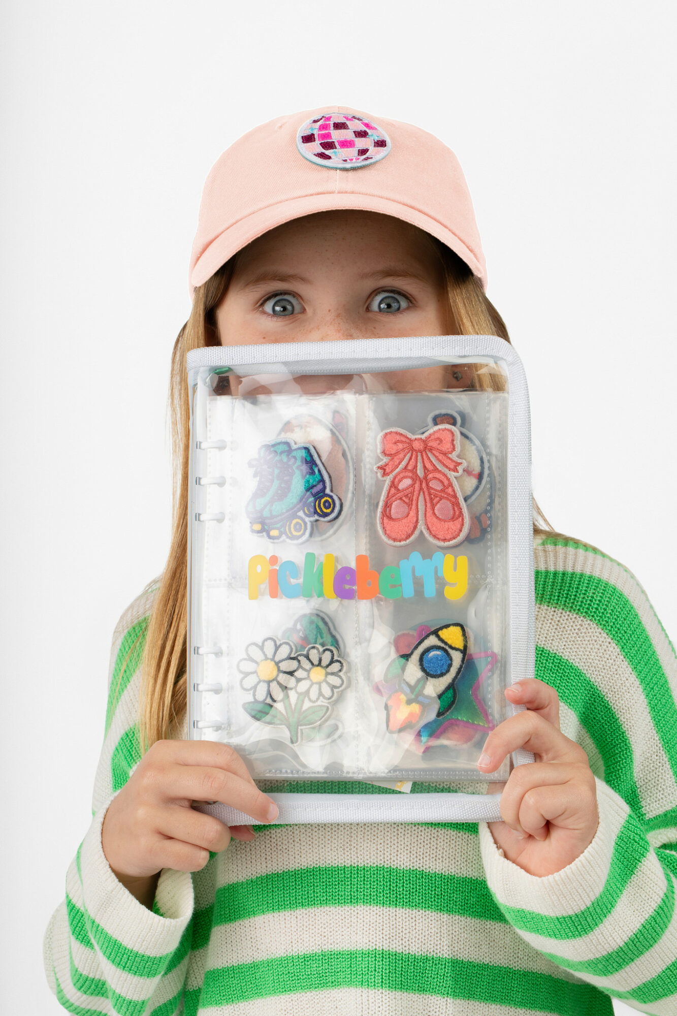 Child wearing a pink cap with a donut patch, holding a clear bag with colorful items, wearing a green and white striped sweater.