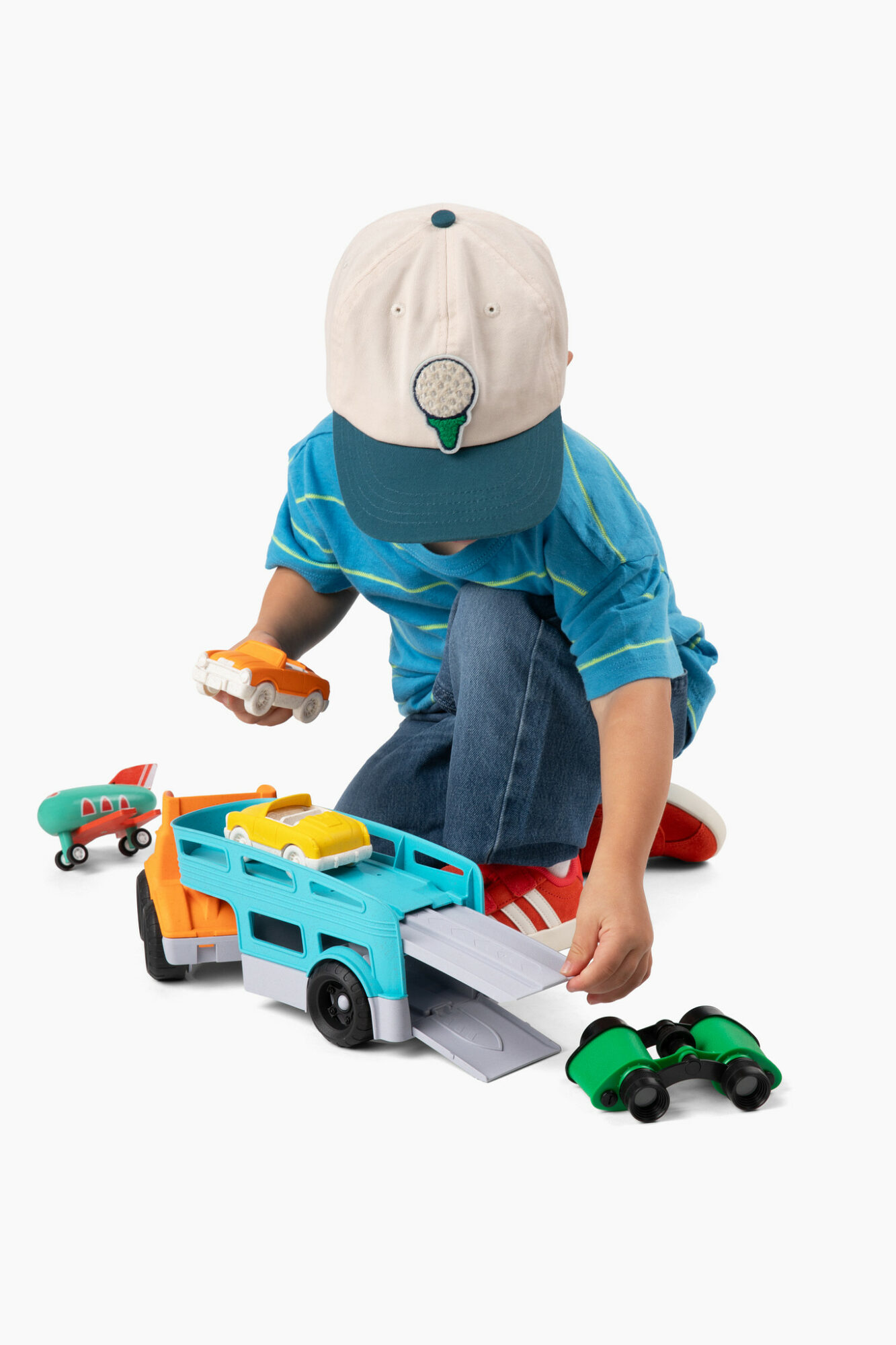 Child playing with toy vehicles, wearing a cap and blue shirt, on a white background.