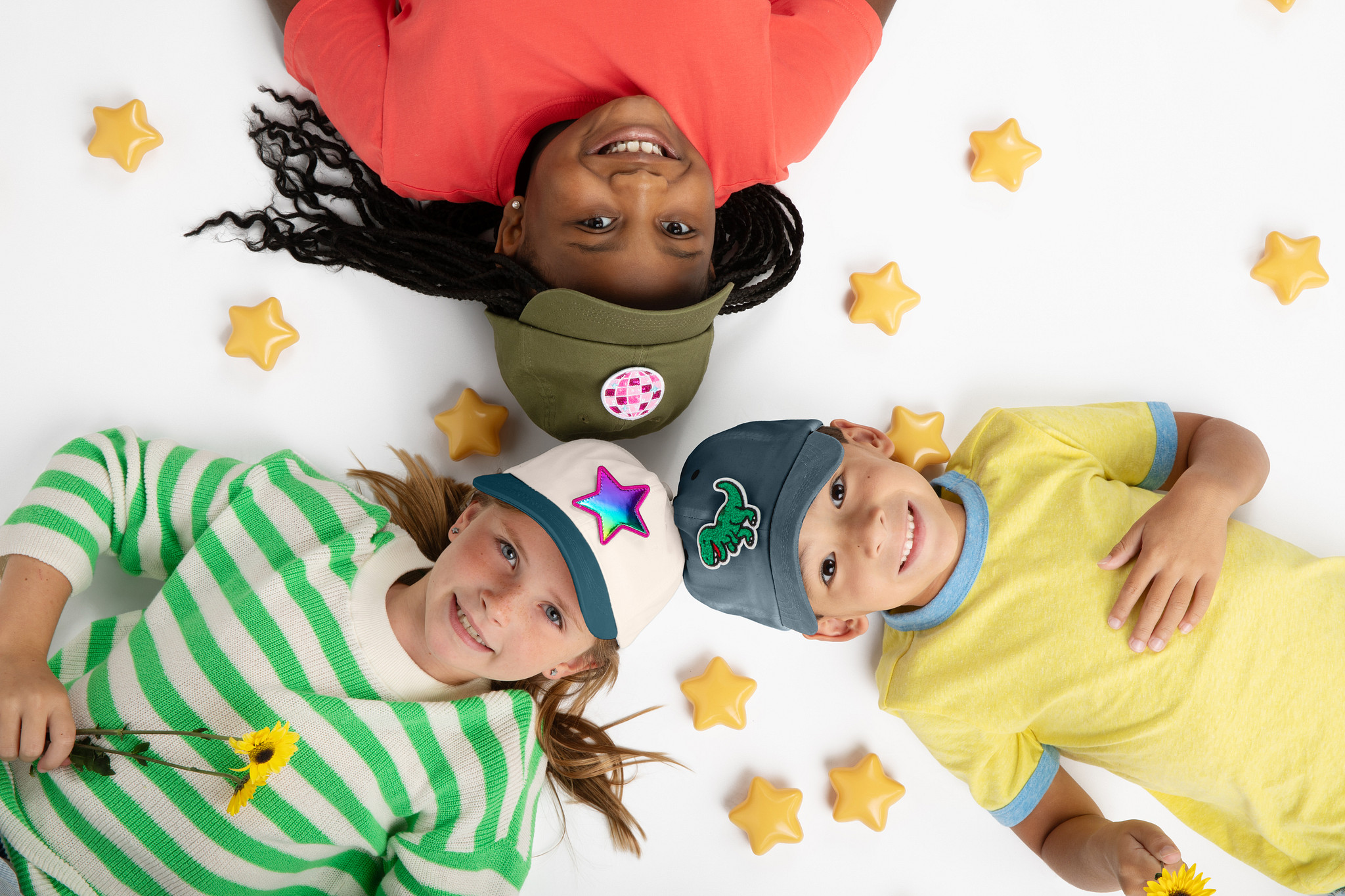 Three children lying on a white surface surrounded by yellow star-shaped objects, smiling and wearing colorful clothing and hats.