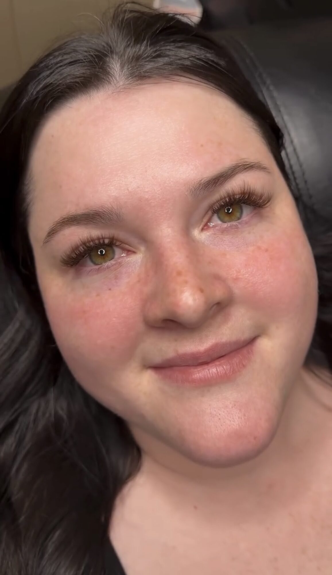 Close-up of a smiling woman with dark hair, light skin, and freckles, looking directly at the camera.