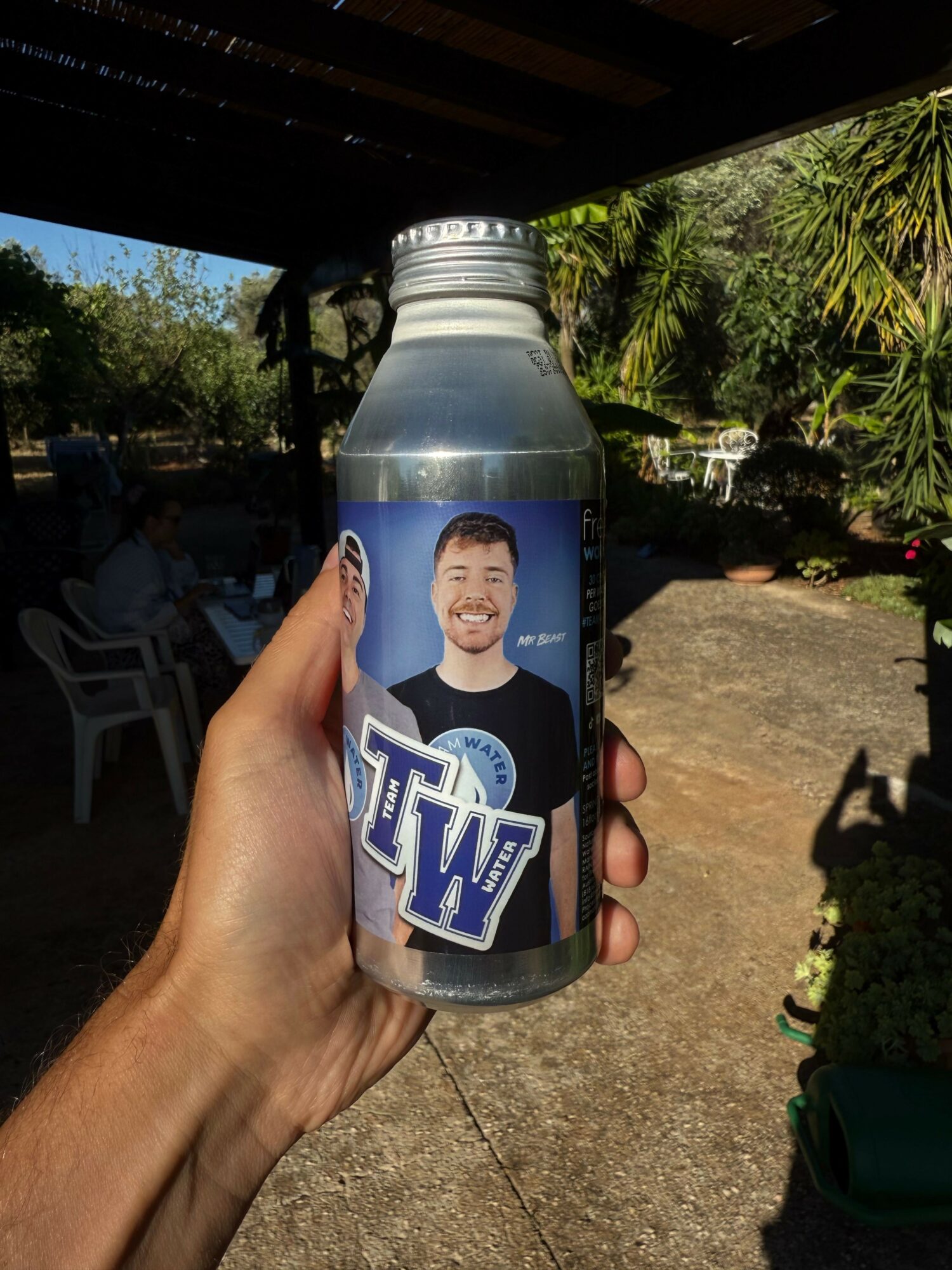 Hand holding a bottle of water outdoors with trees and a pathway in the background.