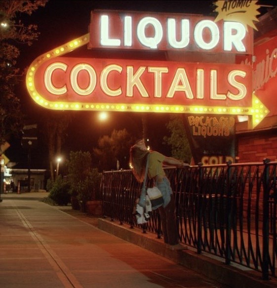 Person leaning on a railing near a brightly lit liquor and cocktails sign at night.