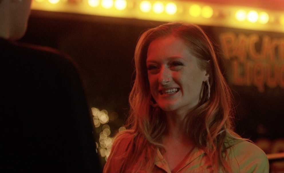 Woman smiling and talking to a person in a dimly lit setting with warm lighting, possibly a bar or club.