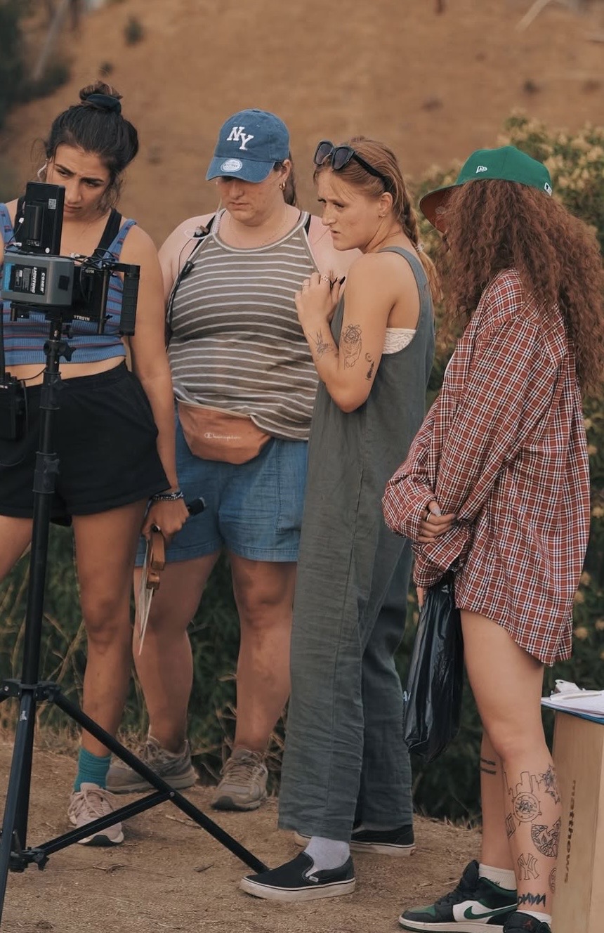 Four women standing outdoors, one with a camera, others looking at it, in casual clothing, with a dirt wall background.