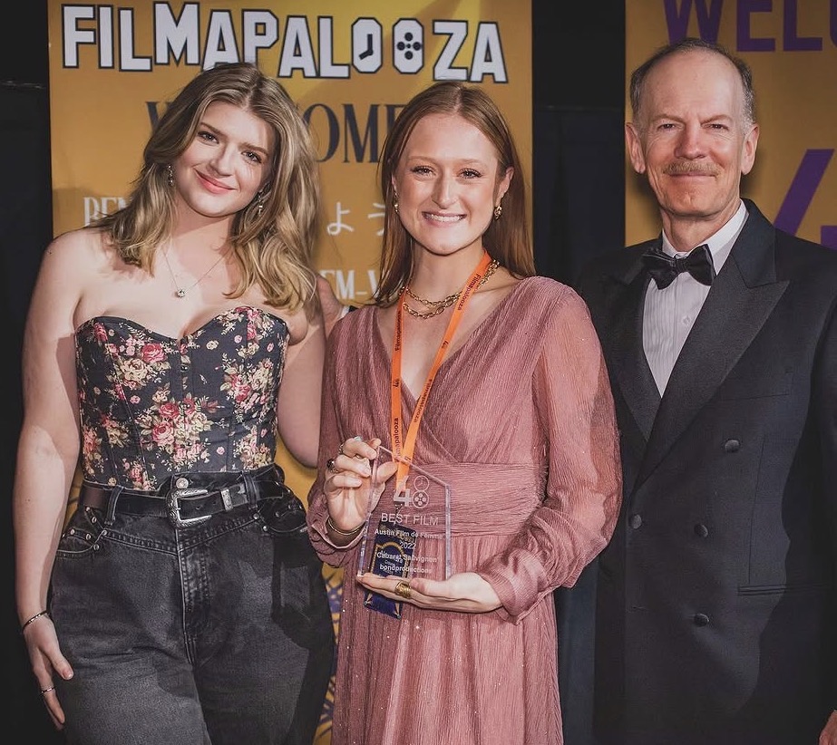 Three people standing together, one holding an award, smiling, in front of a yellow background with text.