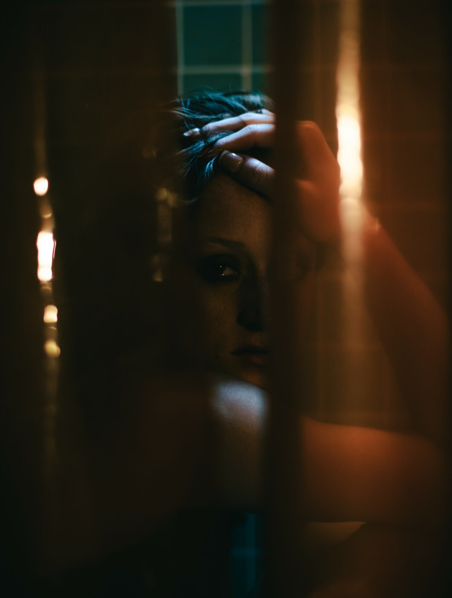 Person with hand on head looking through a window with vertical bars, dim lighting, and warm background glow.