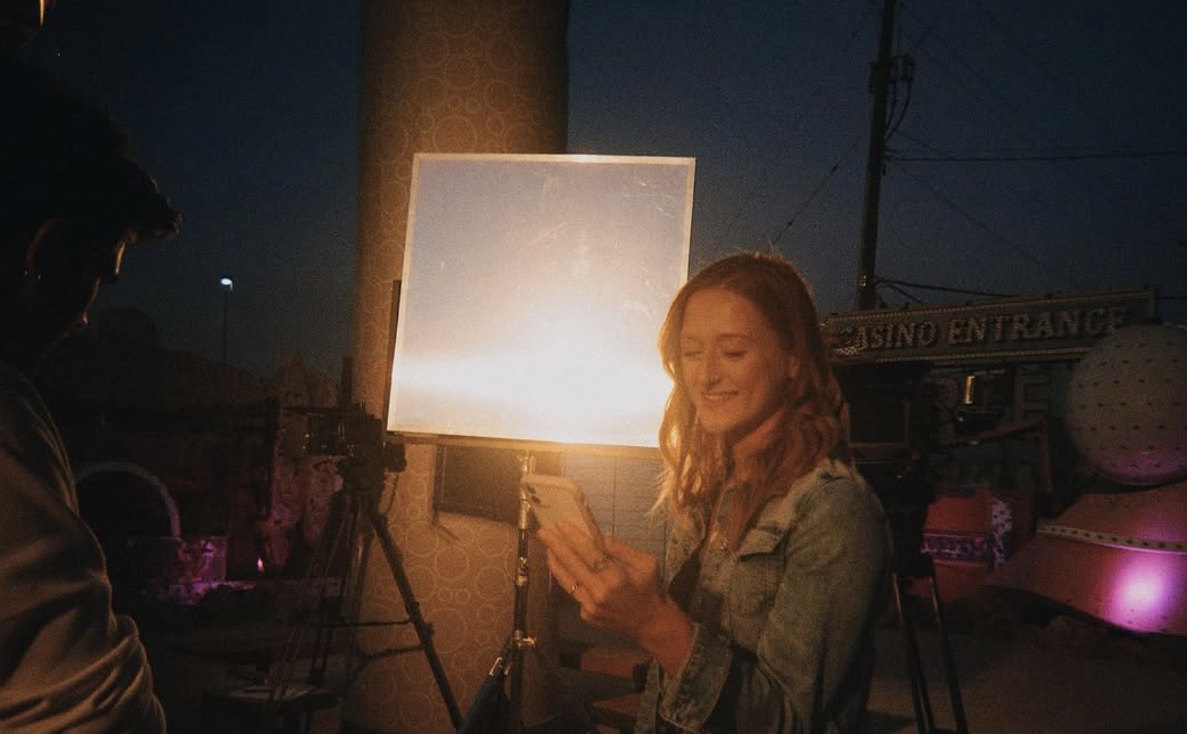 Woman smiling at her phone outdoors at night, illuminated by a bright light, with a dark sky and structures in background.