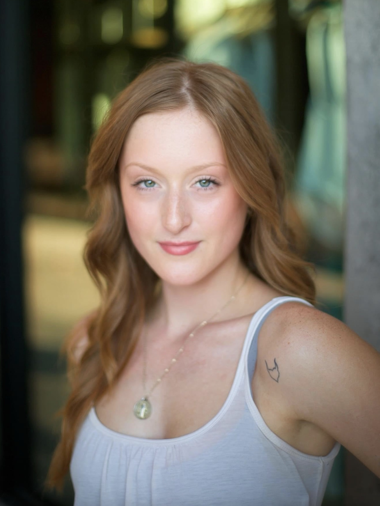 Young woman with long, wavy red hair, wearing a white tank top and a necklace, outdoors with blurred background.
