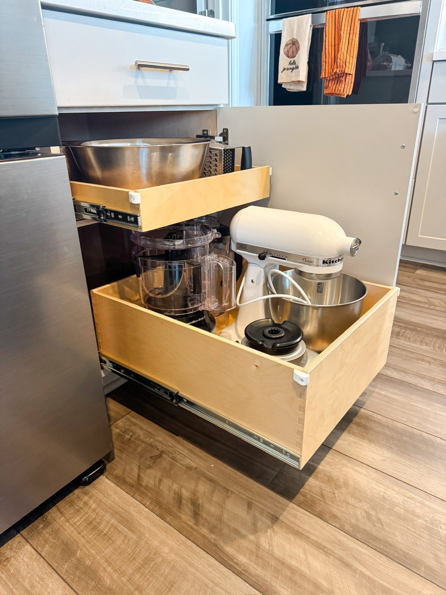 Open kitchen drawer with mixing bowls, glass jars, and a stand mixer inside, next to a refrigerator and kitchen counter.