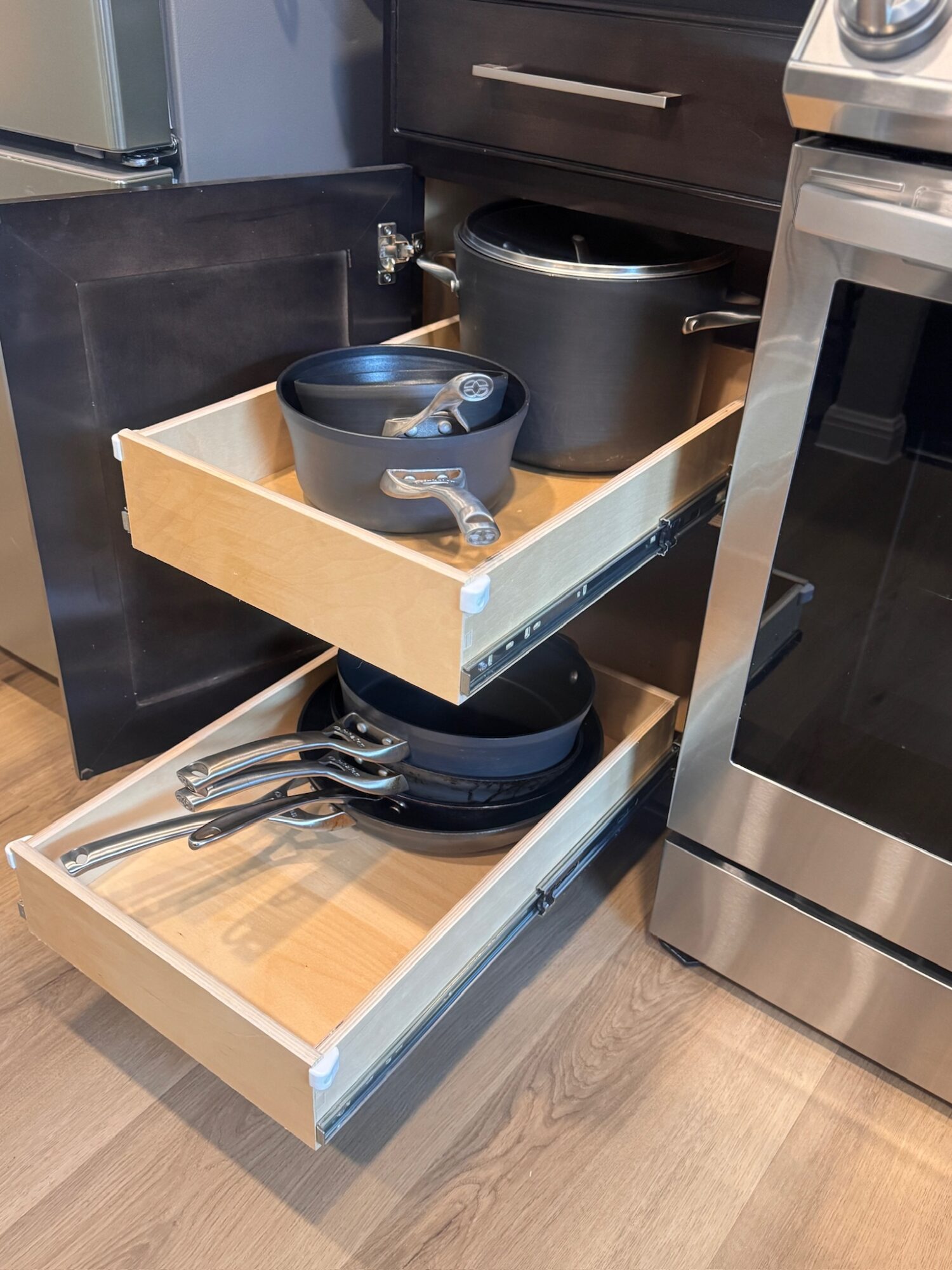 Open kitchen drawer with pots, pans, and utensils inside, next to an oven and a trash bin.