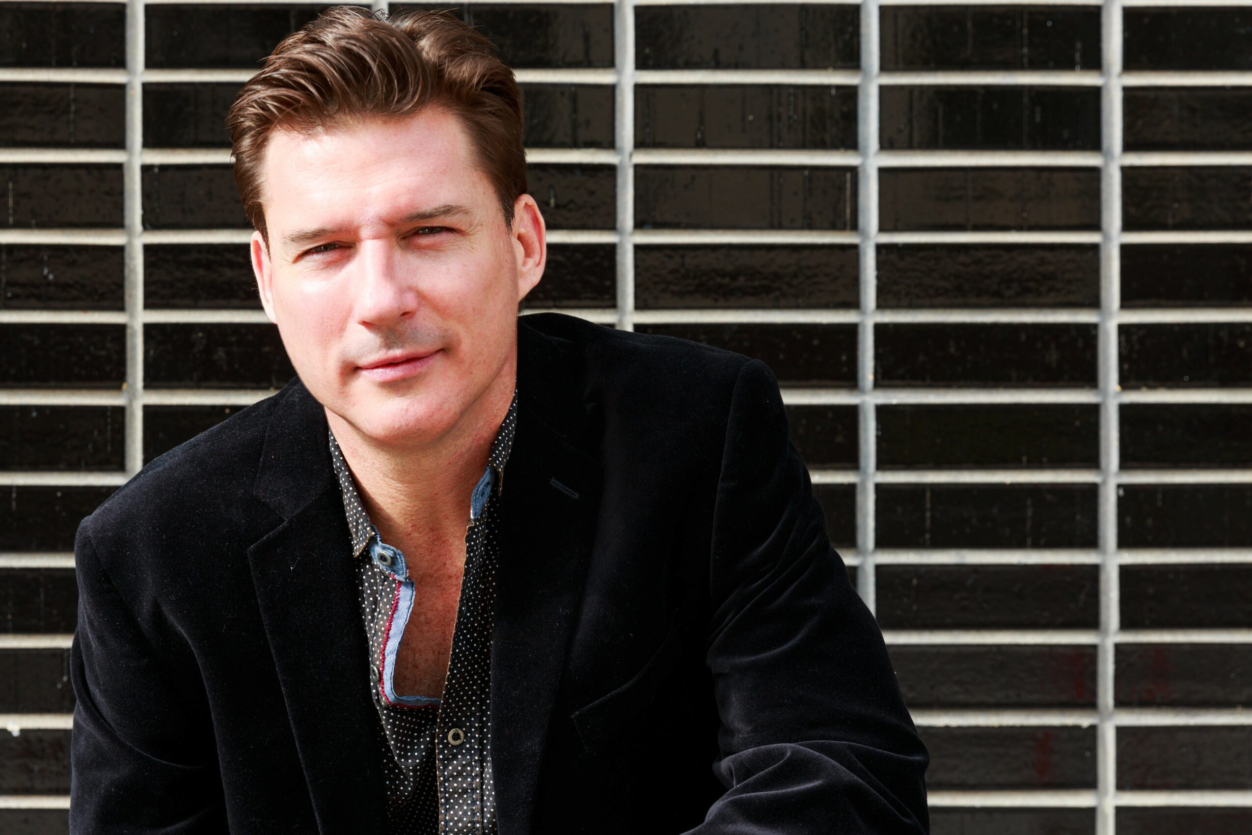 Young man with brown hair wearing a black jacket in front of a grid background.