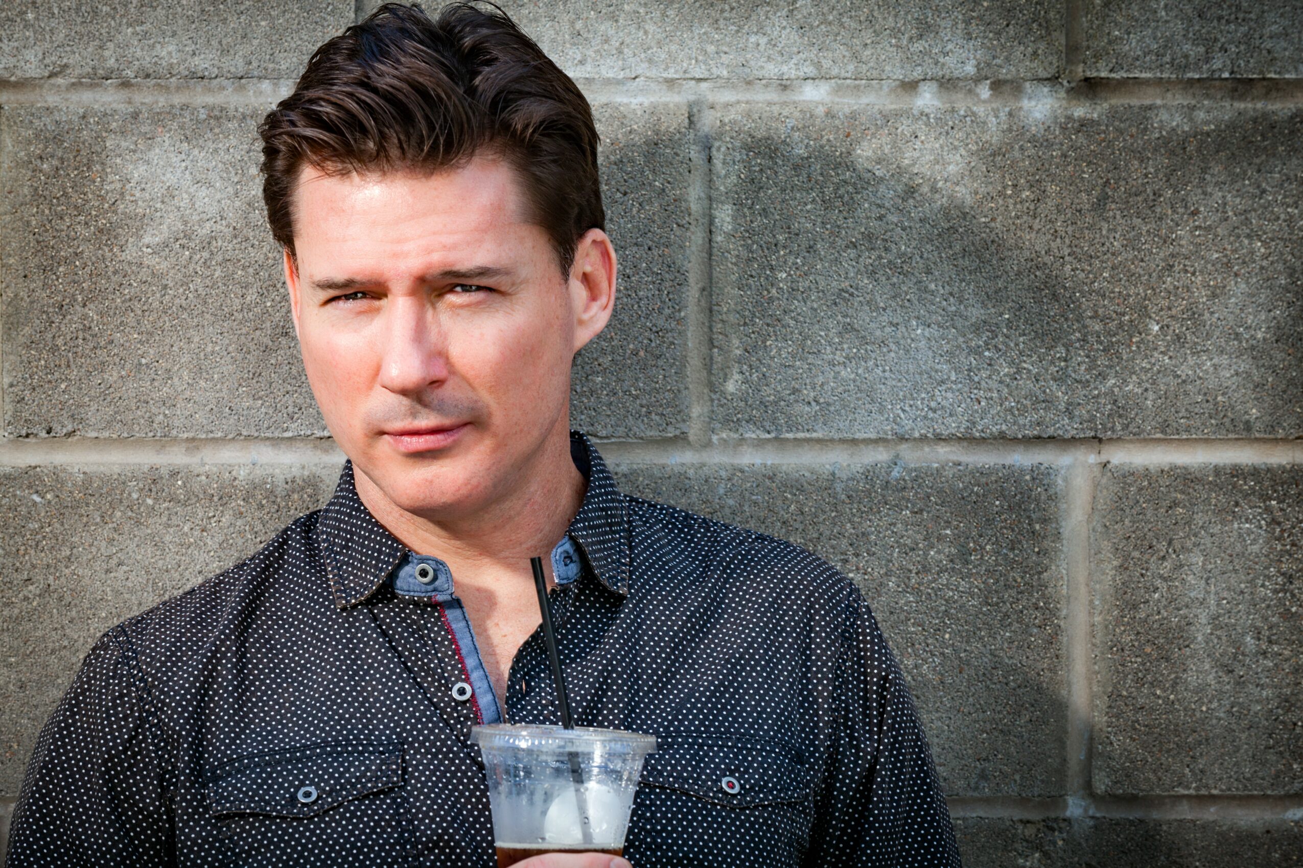Young man with dark hair and a serious expression stands against a gray brick wall, wearing a dark patterned shirt.