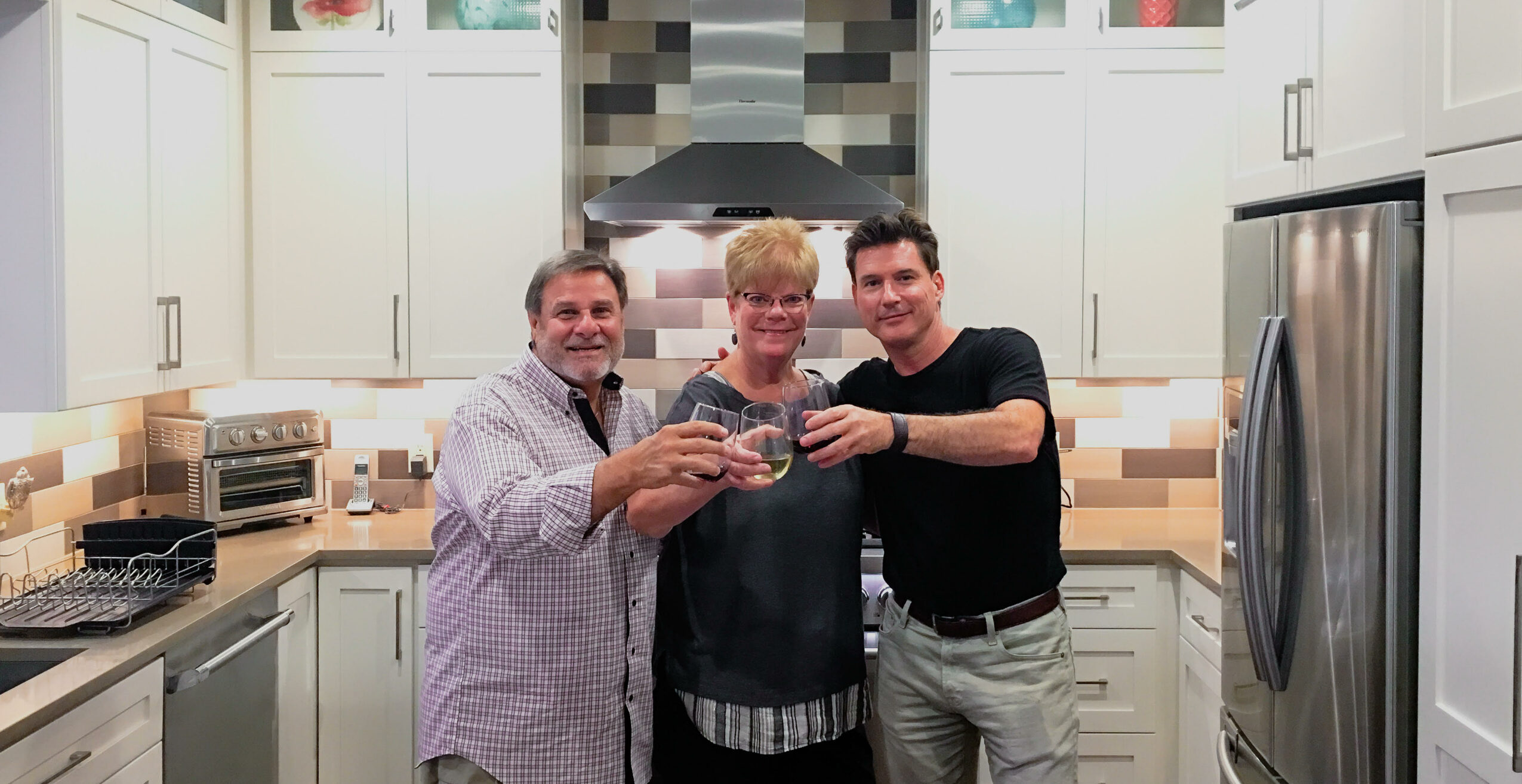 Three people smiling and holding a glass in a kitchen with white cabinets and stainless steel appliances.
