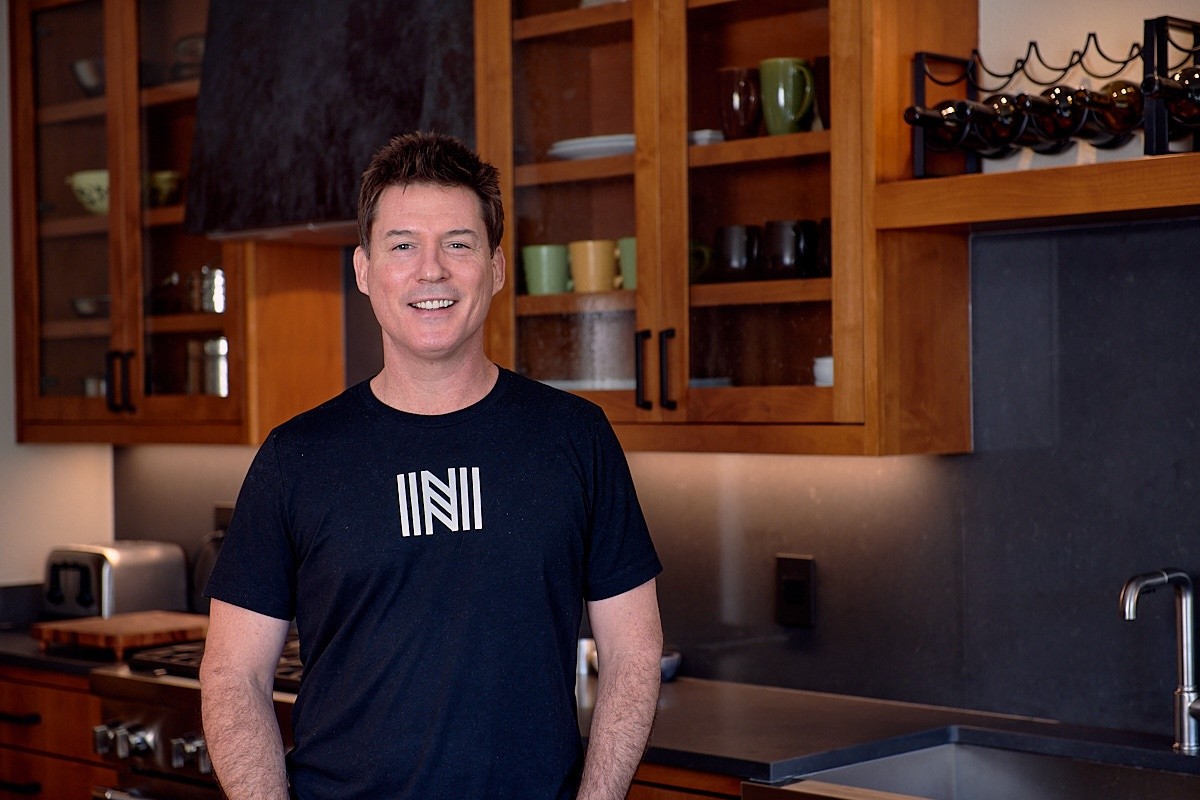 Young man with short hair smiling in a kitchen with wooden cabinets and black countertops.
