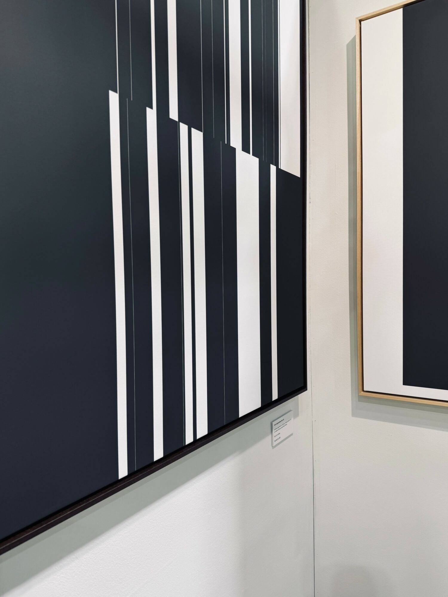 Abstract artwork with black, white, and gray vertical stripes on a wall, framed in gold, in a gallery setting.