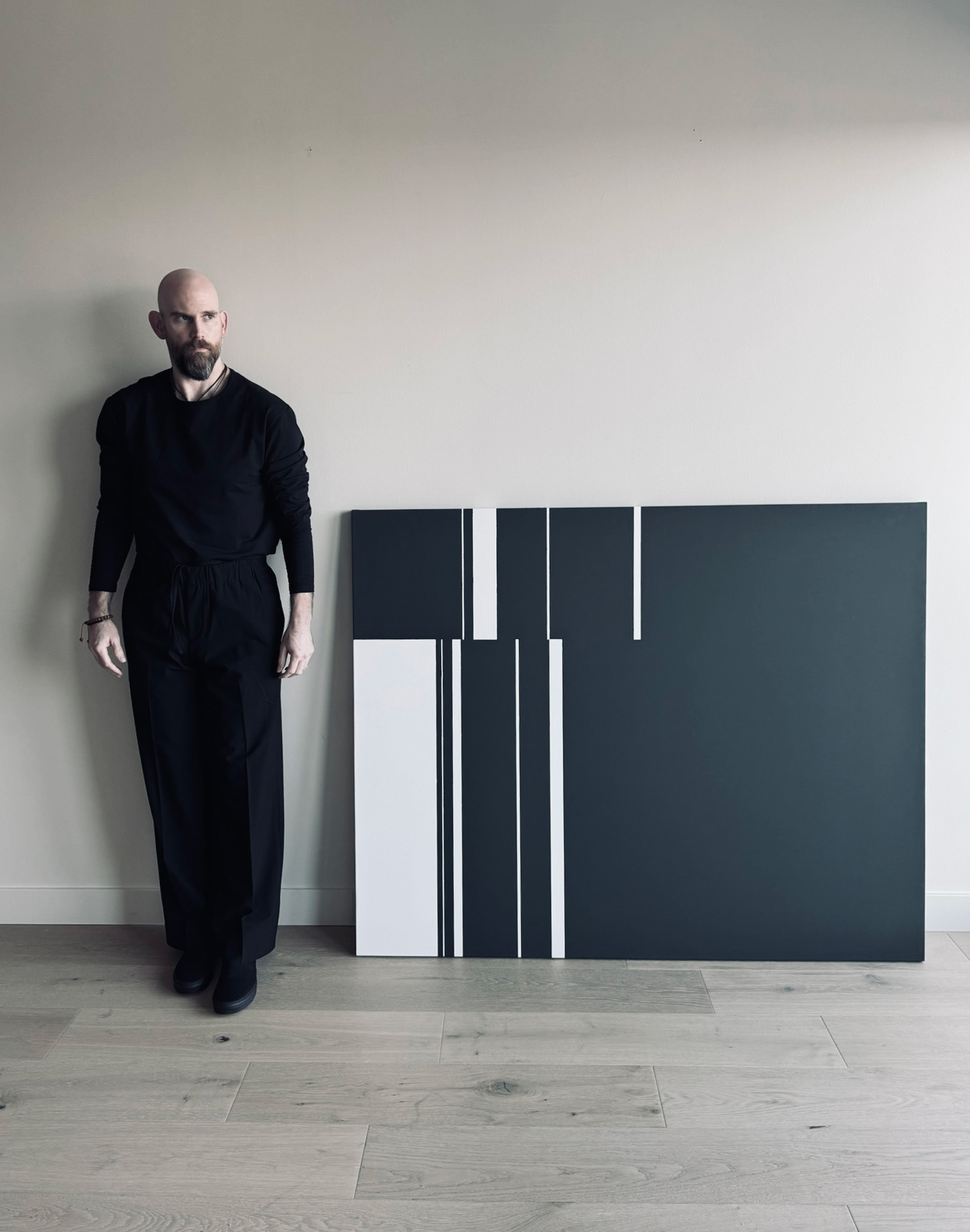 Man with a beard in black clothing standing next to abstract black and white artwork on a wall, with light-colored floor.