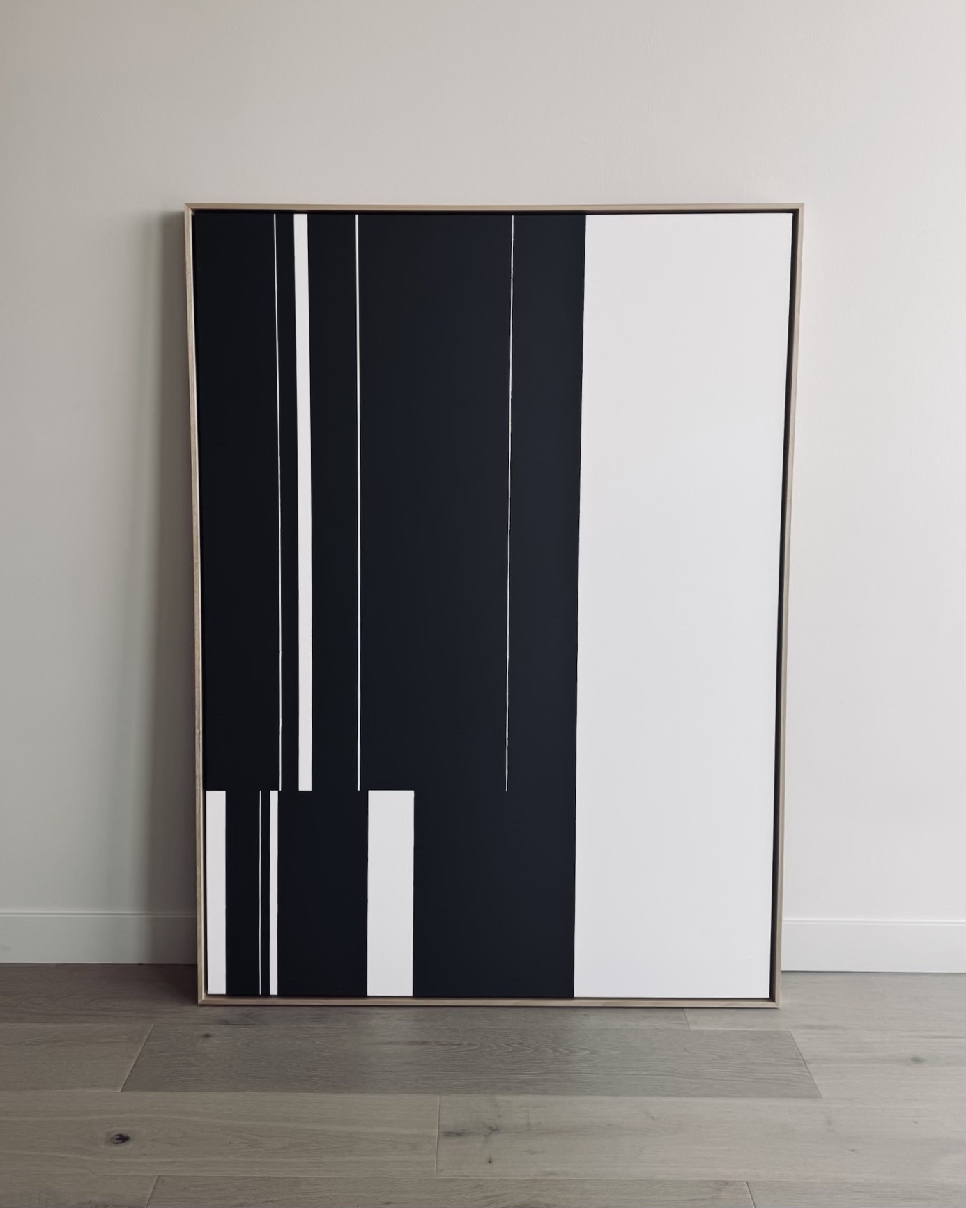 Abstract black and white geometric artwork leaning against a wall, with vertical lines and blocks, on a wooden floor.
