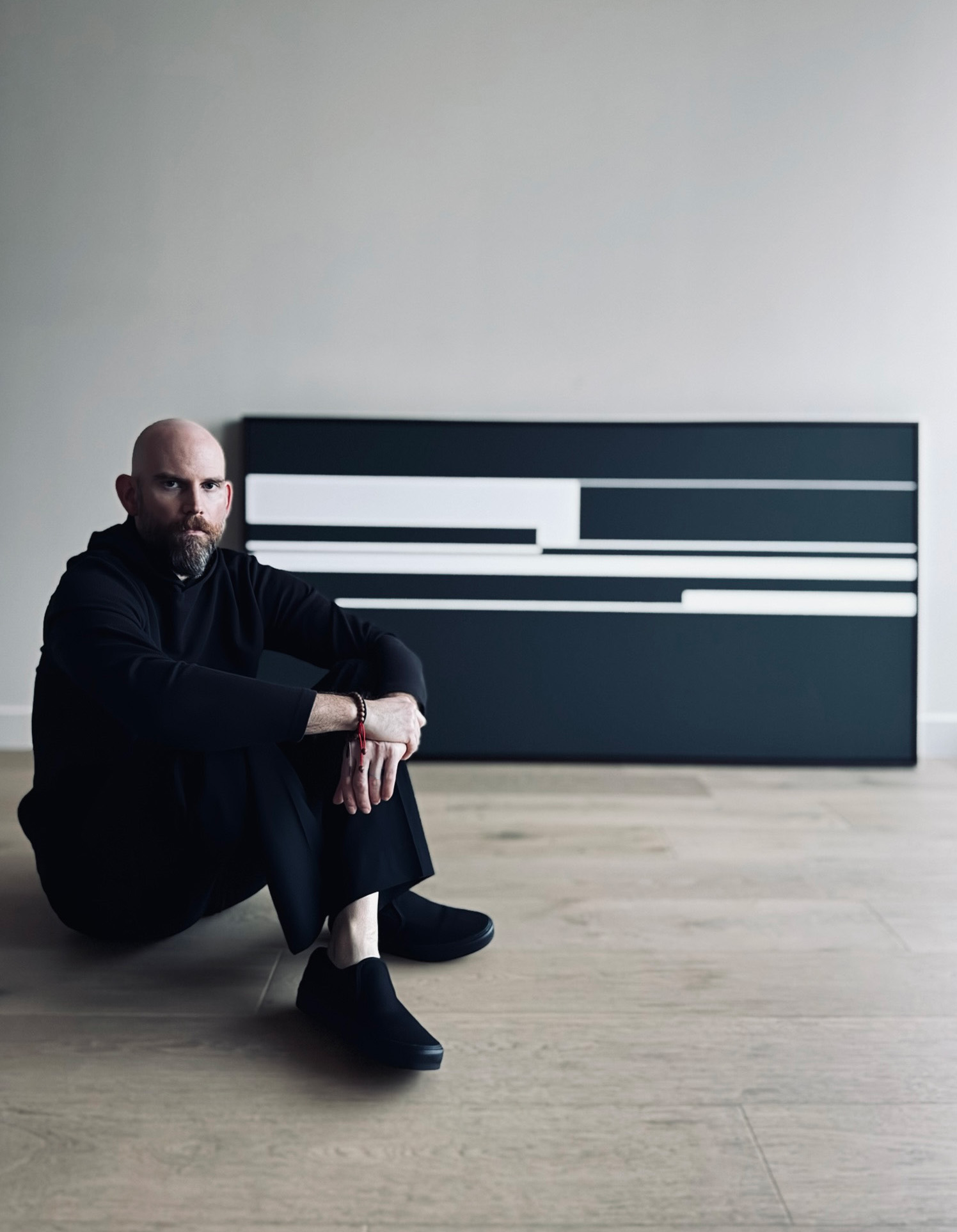 Man sitting on the floor in front of a large abstract black and white artwork, with a plain wall and wooden floor.