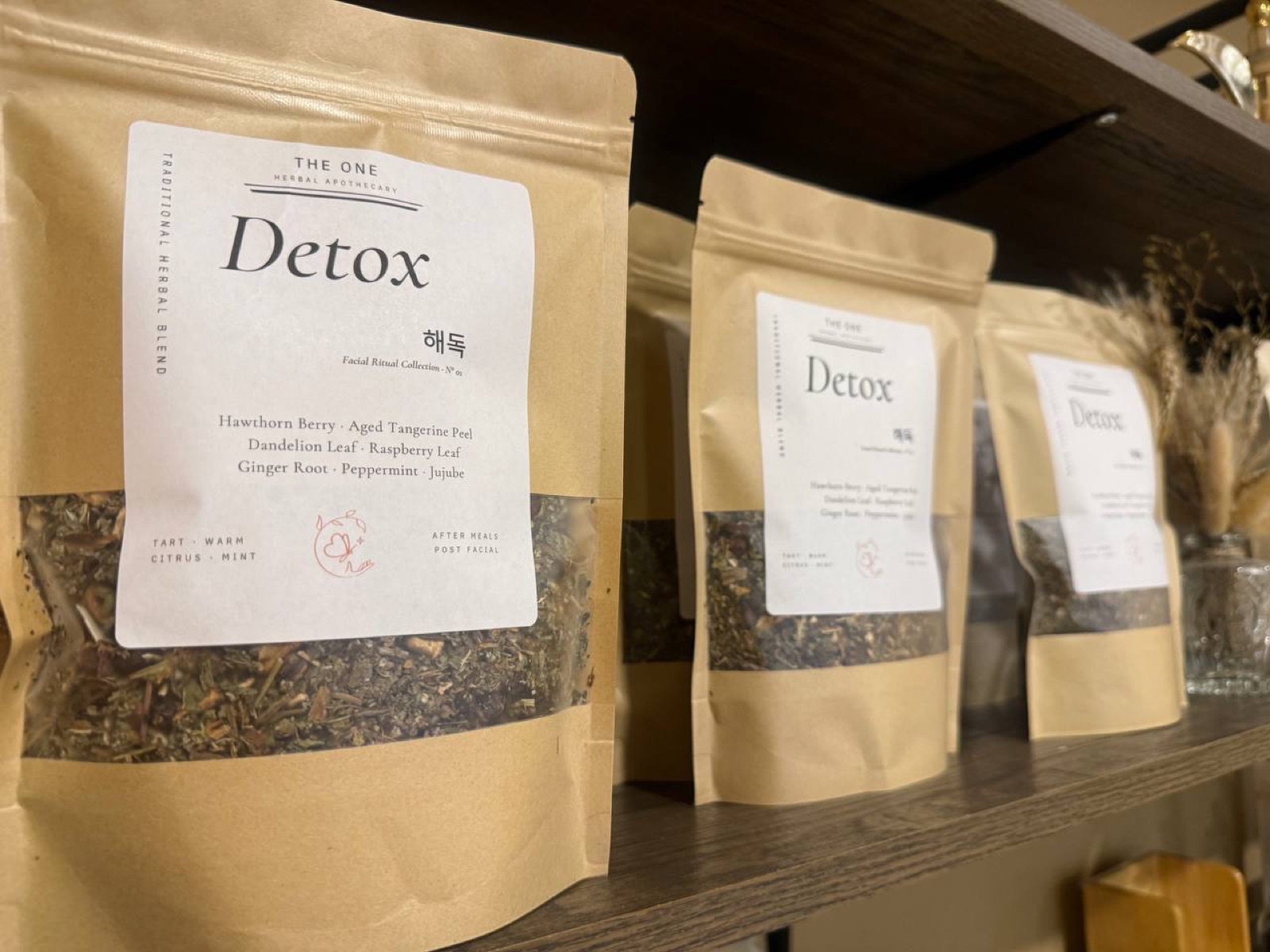Three beige paper bags labeled 'Detox' on a wooden shelf, containing herbal tea blends, with a plant in the background.
