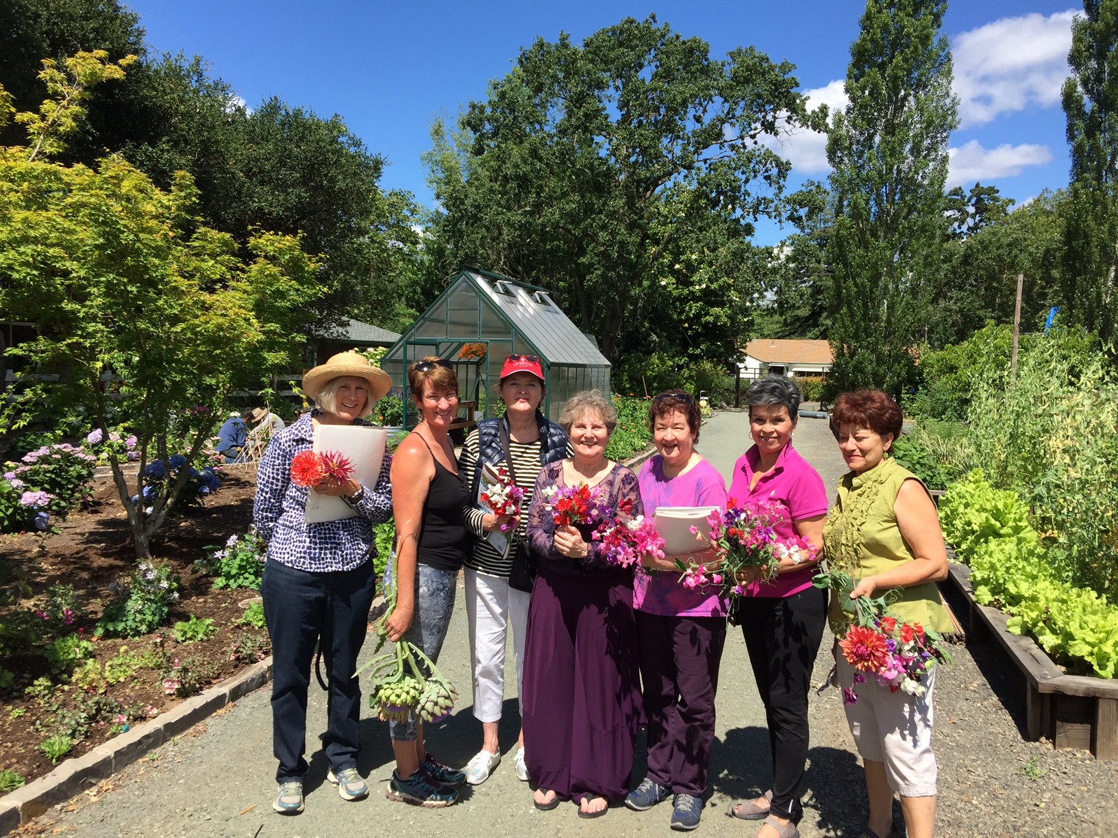 Group of seven women standing outdoors in a garden, holding flowers and smiling, with trees and a small building in the background.