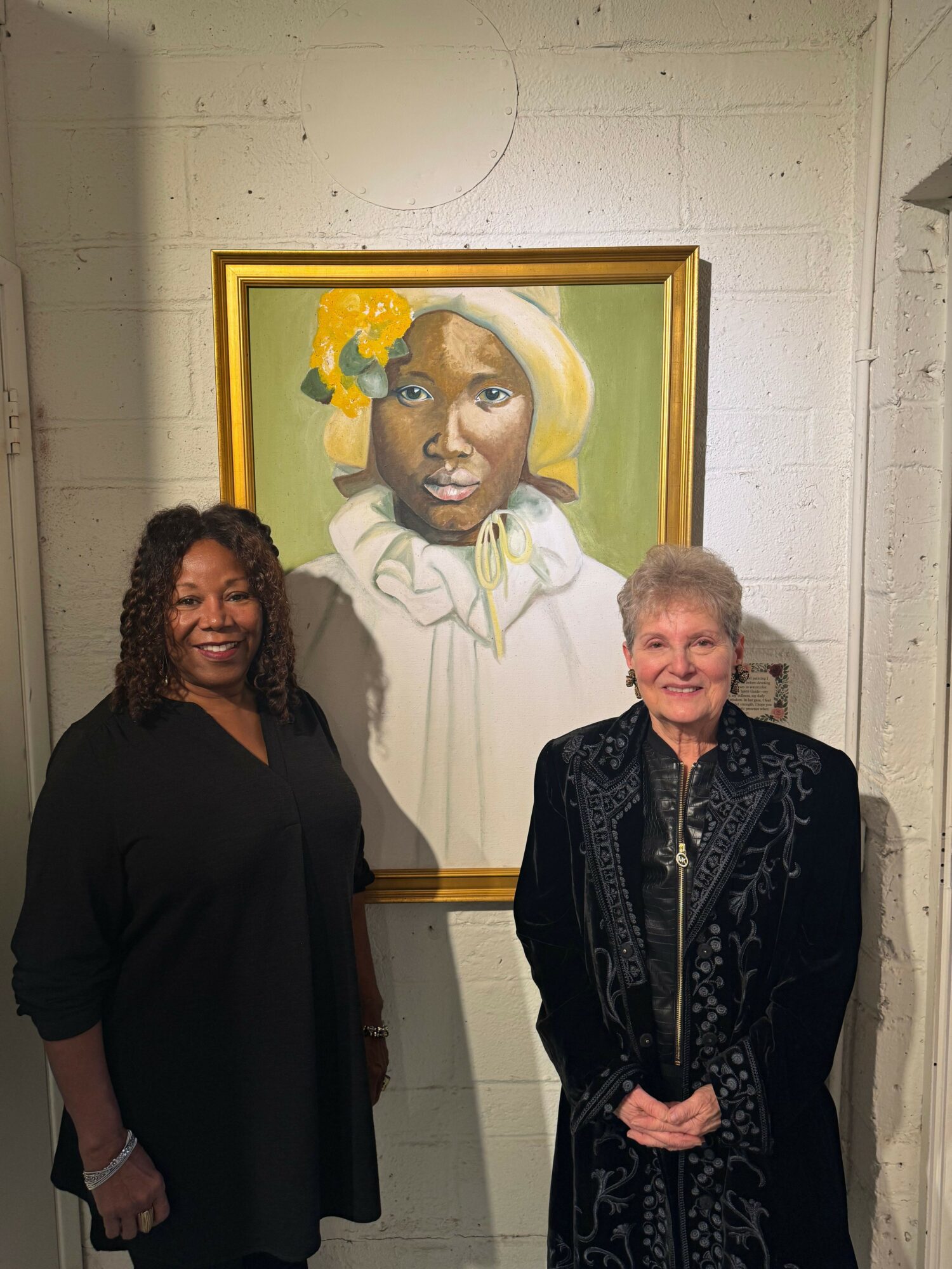 Two women standing in front of a portrait painting on a white wall, one with short gray hair and the other with long dark hair.