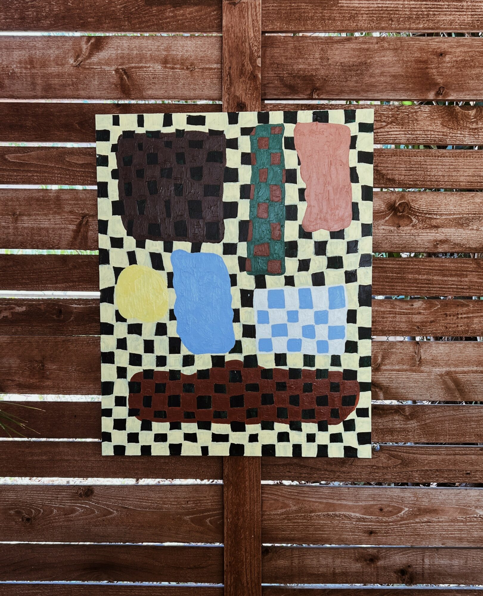 Abstract artwork with colorful shapes on a checkered background, mounted on a wooden fence.