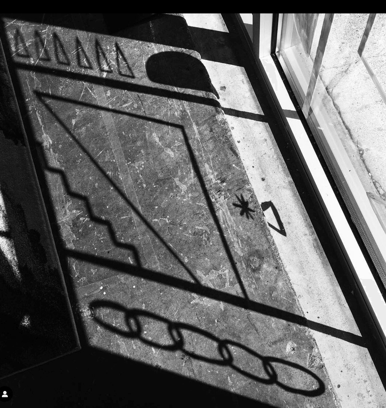Shadow of a window frame and objects cast on a concrete floor, with a window and wall visible. Black and white.