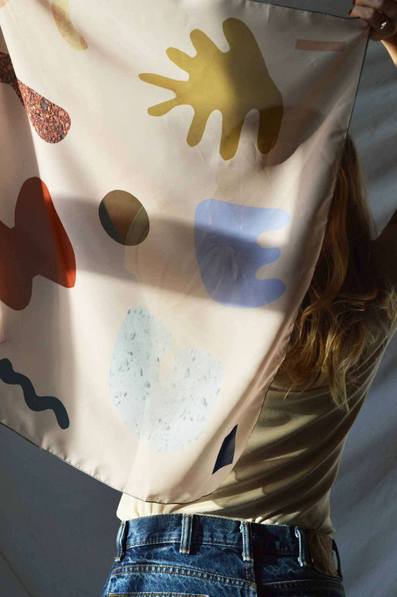 Person holding a fabric with colorful hand and shape patterns, partially covering their face, wearing a beige top and blue jeans.