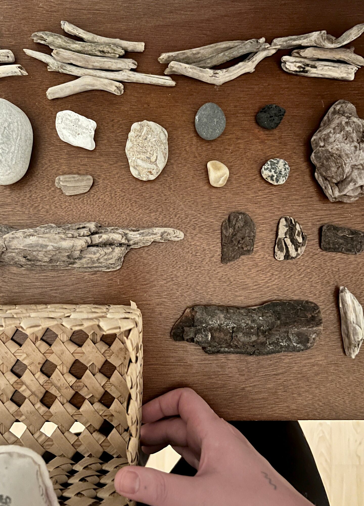 Collection of various rocks and driftwood pieces on a wooden surface, with a hand holding a woven basket in the bottom left.