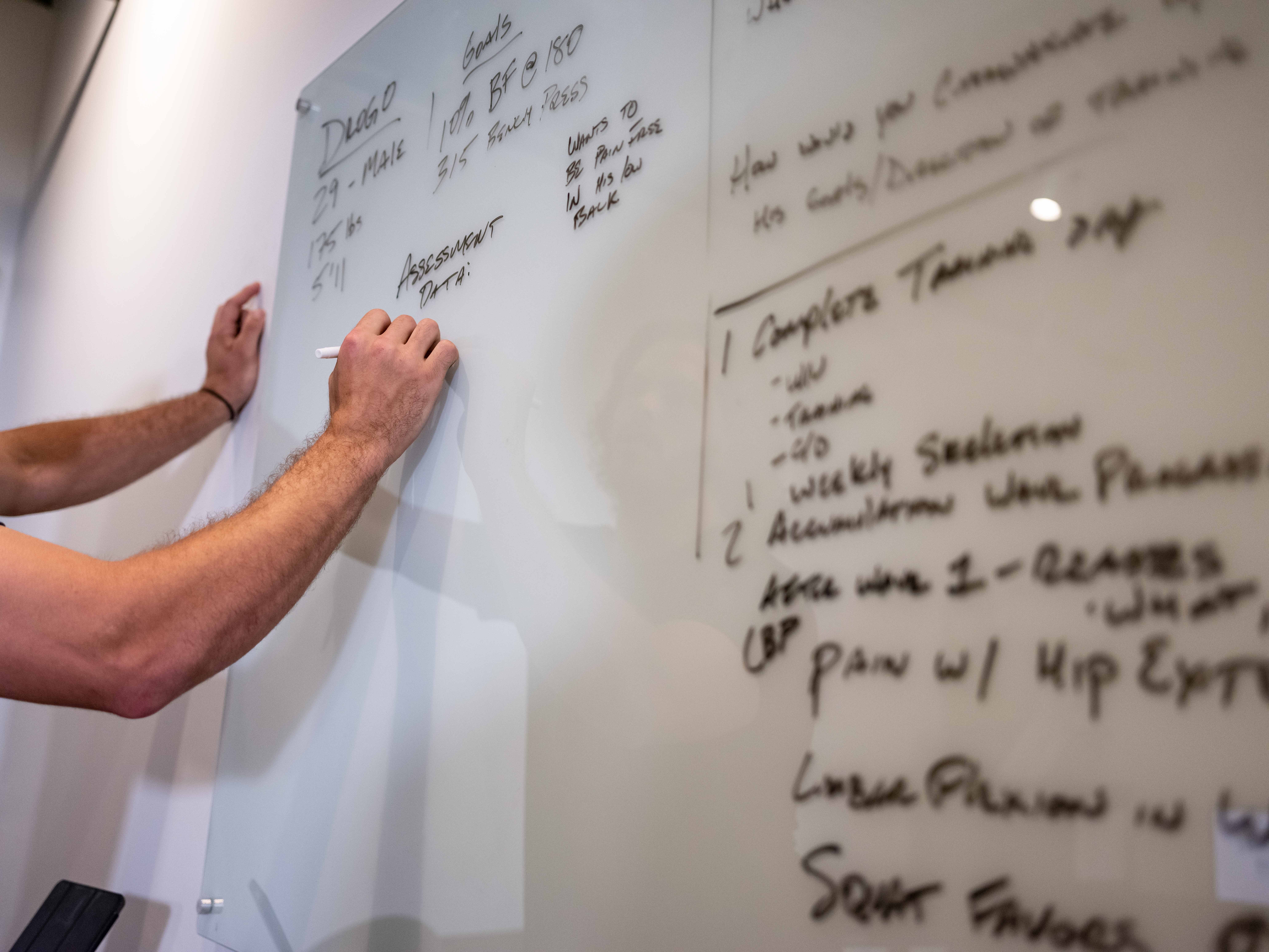 Person writing on a whiteboard with handwritten notes and lists, partially visible arm and hand, background of a wall.