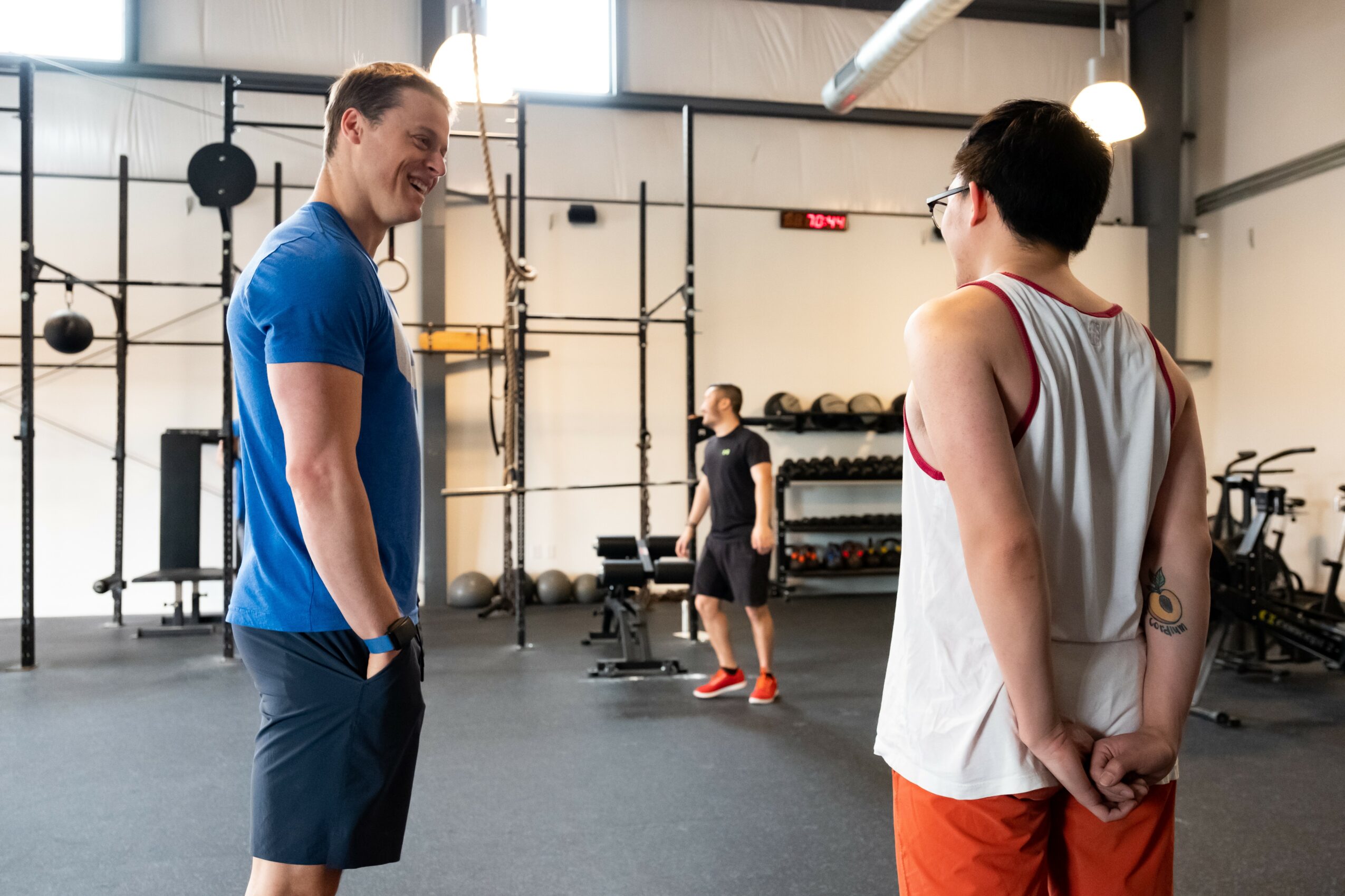 Two men talking in a gym, one in a blue shirt and the other in a white tank top with red accents, with gym equipment in background.