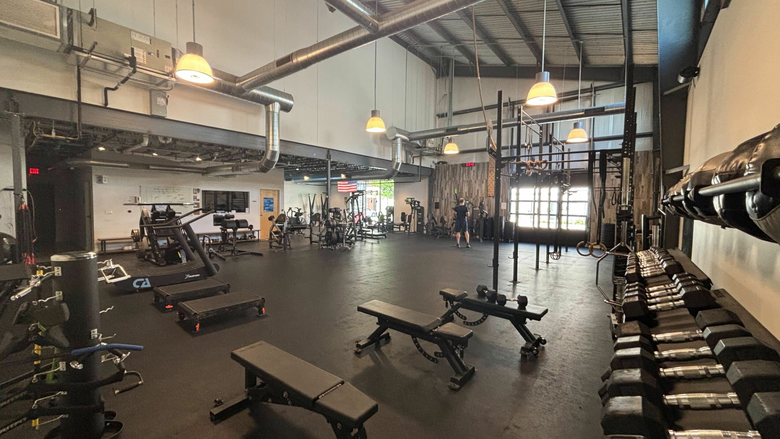 Empty gym with workout benches, weights, and exercise equipment, illuminated by hanging lights and large windows.