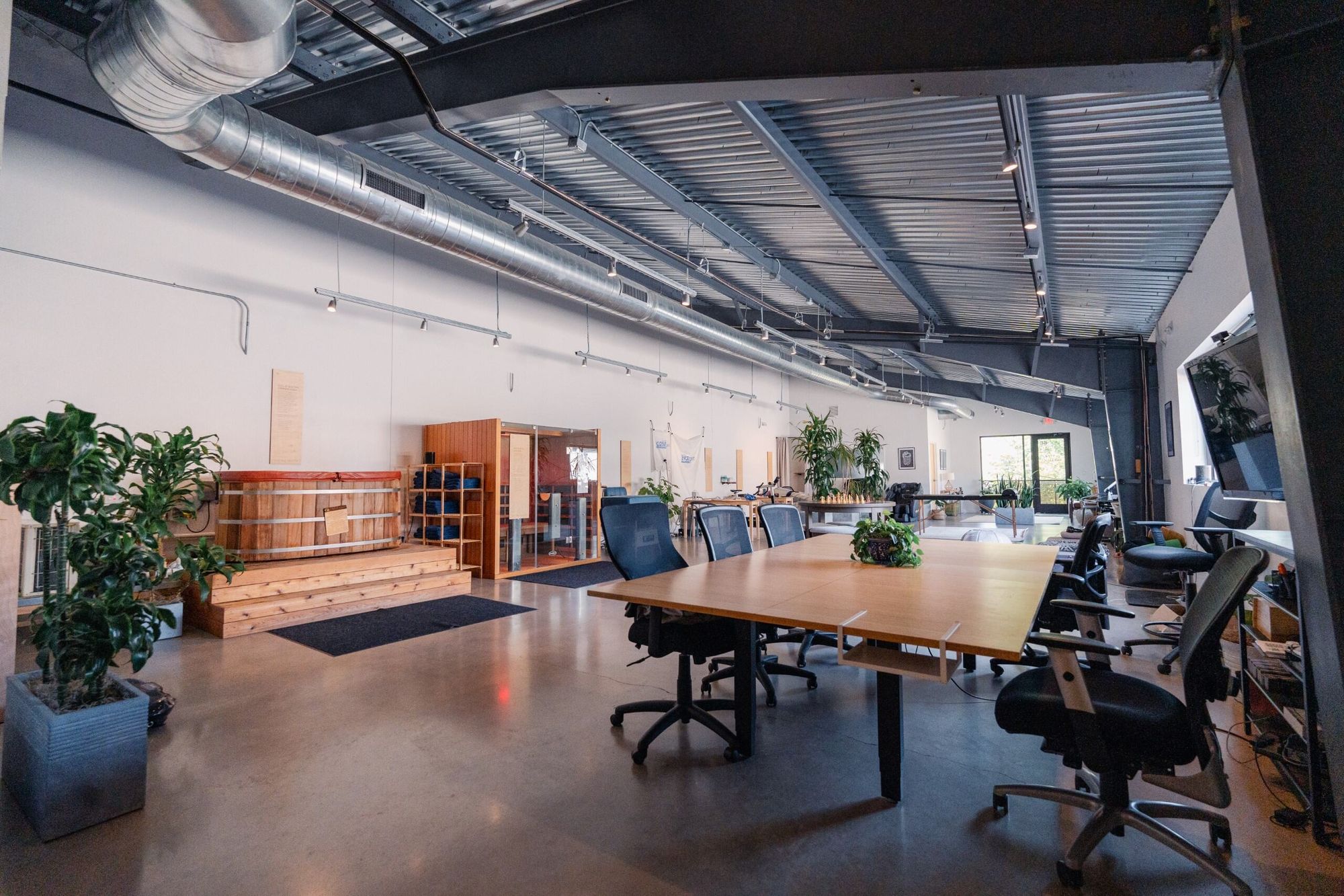 Modern office space with large table, chairs, plants, and industrial ceiling, viewed from the entrance.
