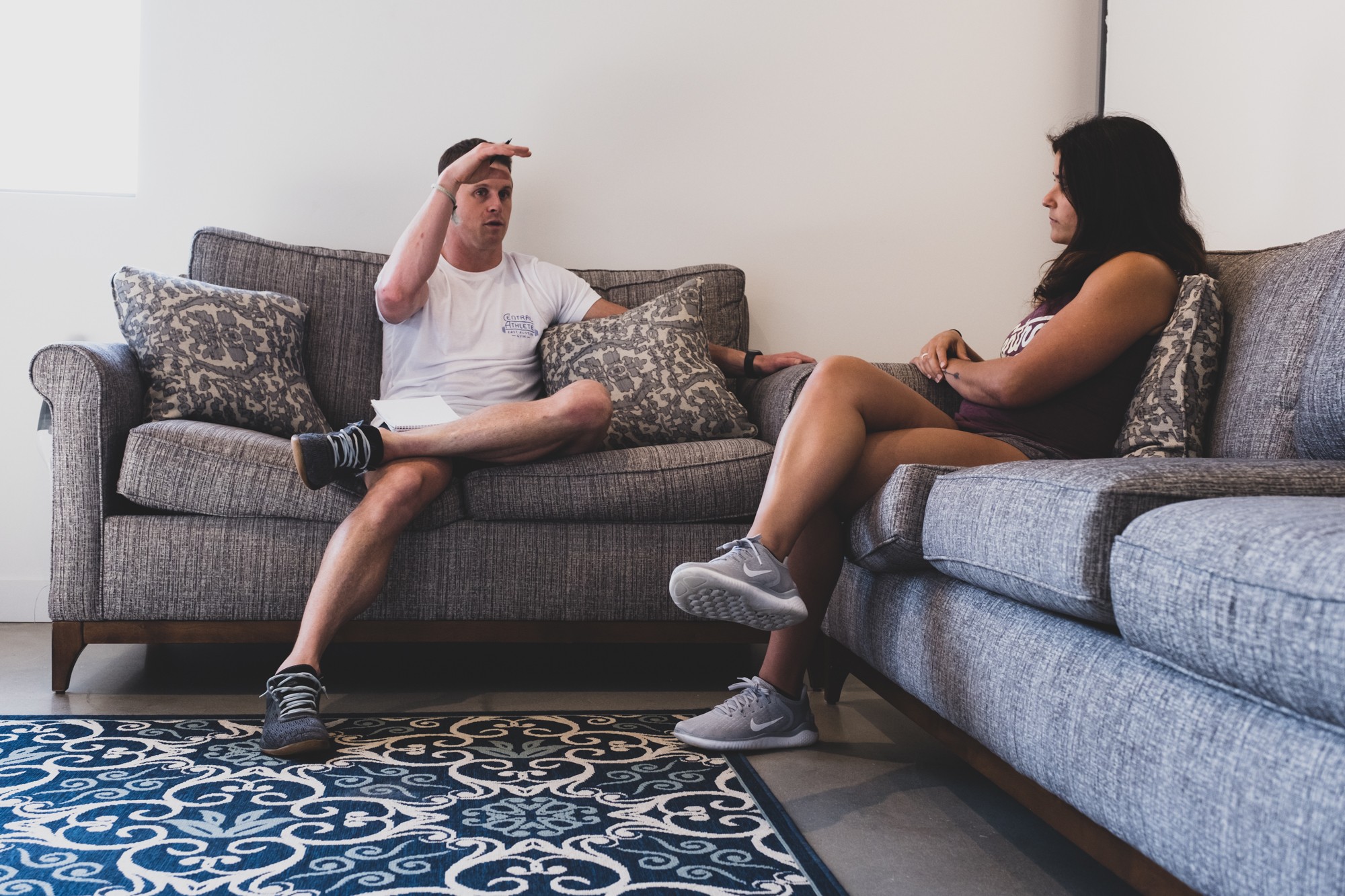 Two people sitting on a gray sofa, talking, with a patterned rug on the floor, in a room with white walls.