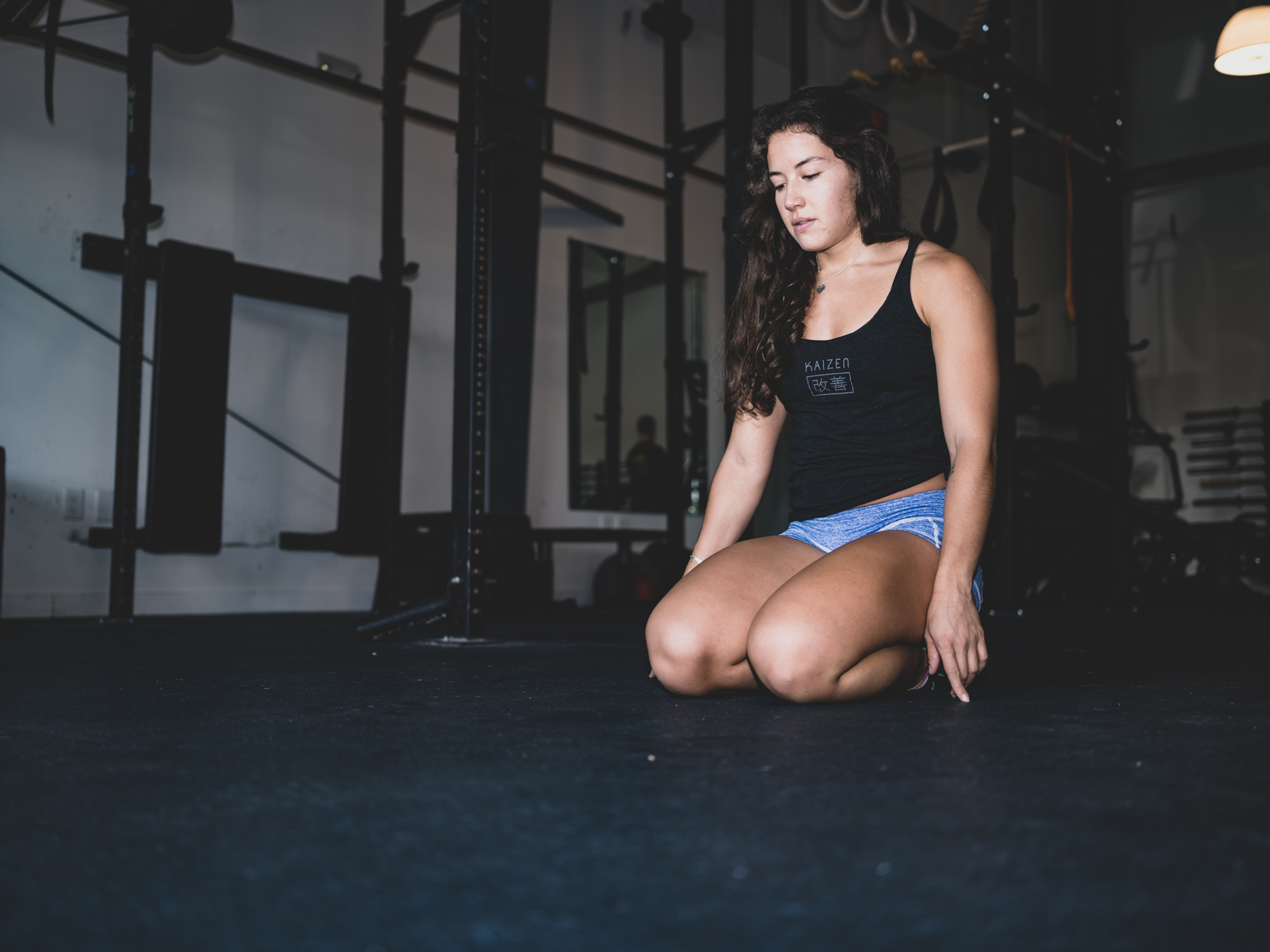 Young woman kneeling on gym floor, looking down, with gym equipment in background.
