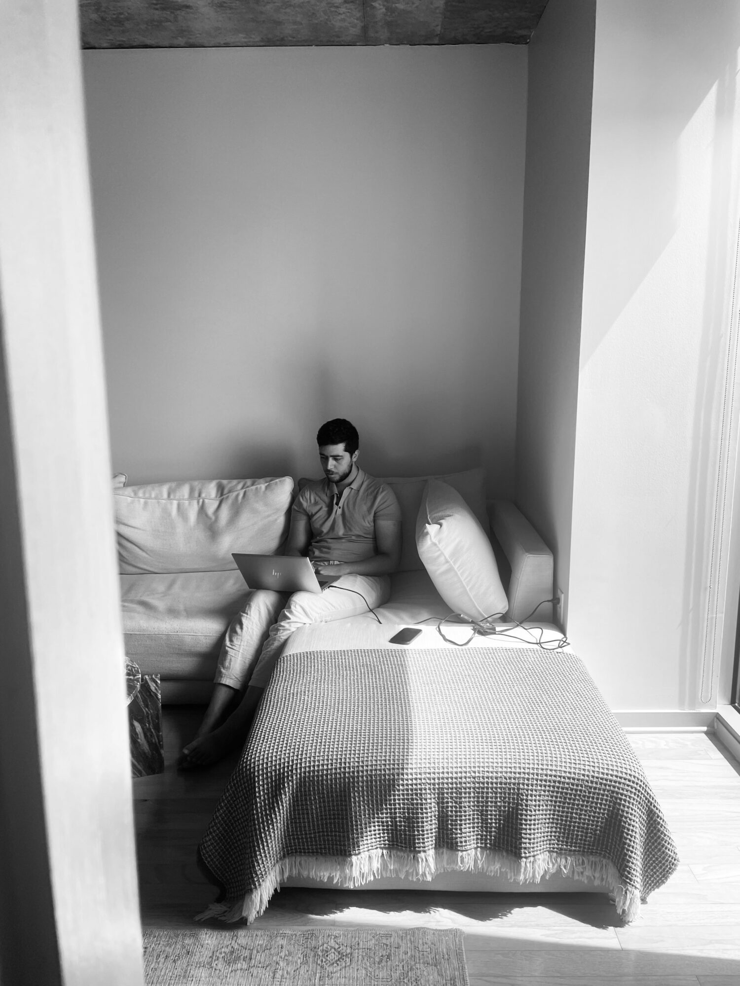 Person sitting on a bed using a laptop in a minimalistic room with plain walls and natural light.