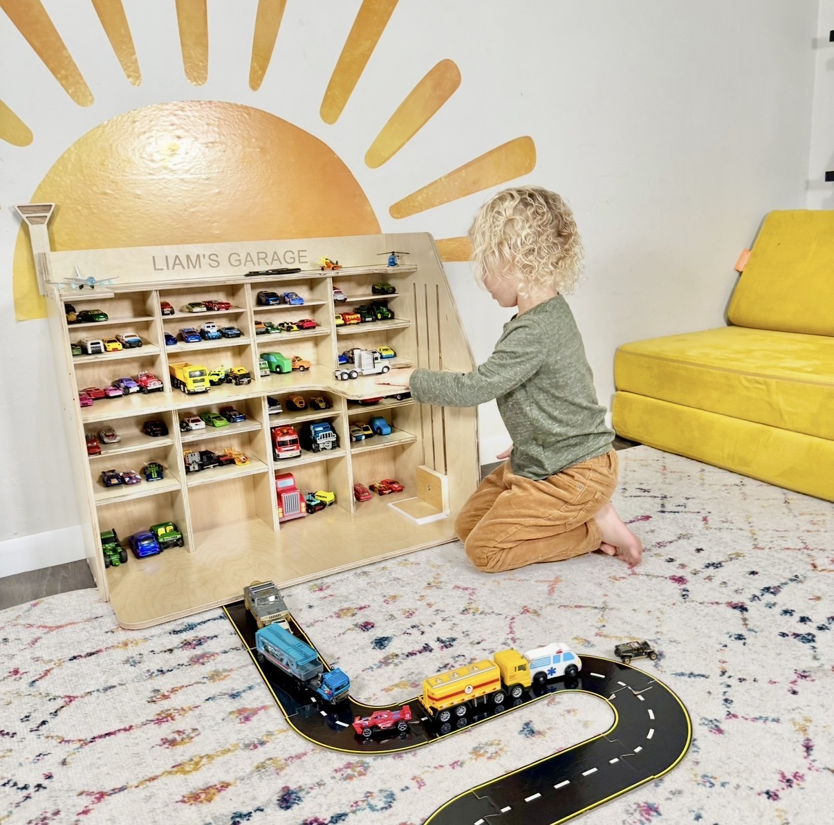Child with curly hair playing with a wooden car garage and toy cars on a colorful rug, with a yellow couch nearby.