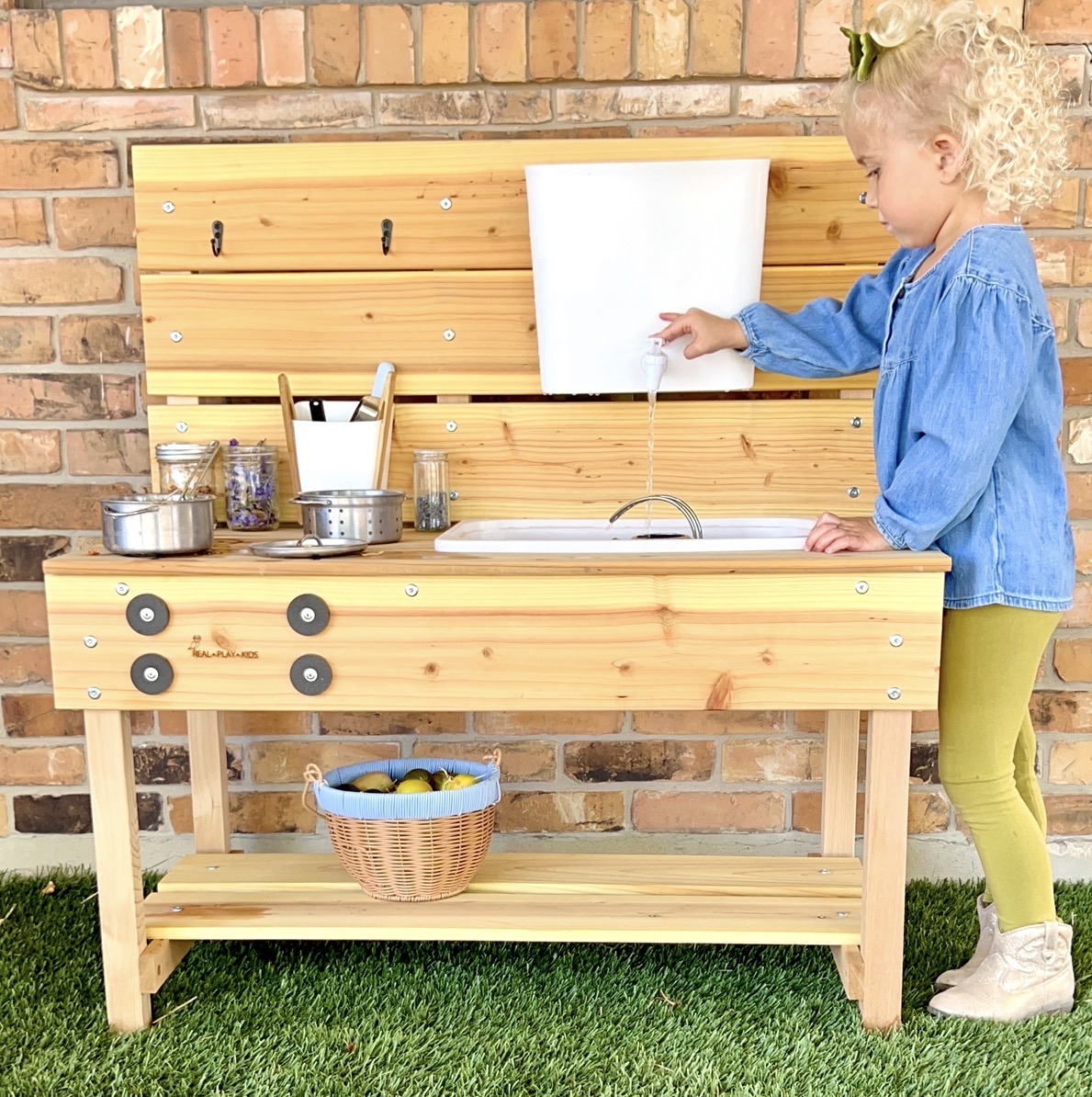 Child playing with water at outdoor wooden play kitchen against brick wall, with basket on lower shelf, grass in foreground.