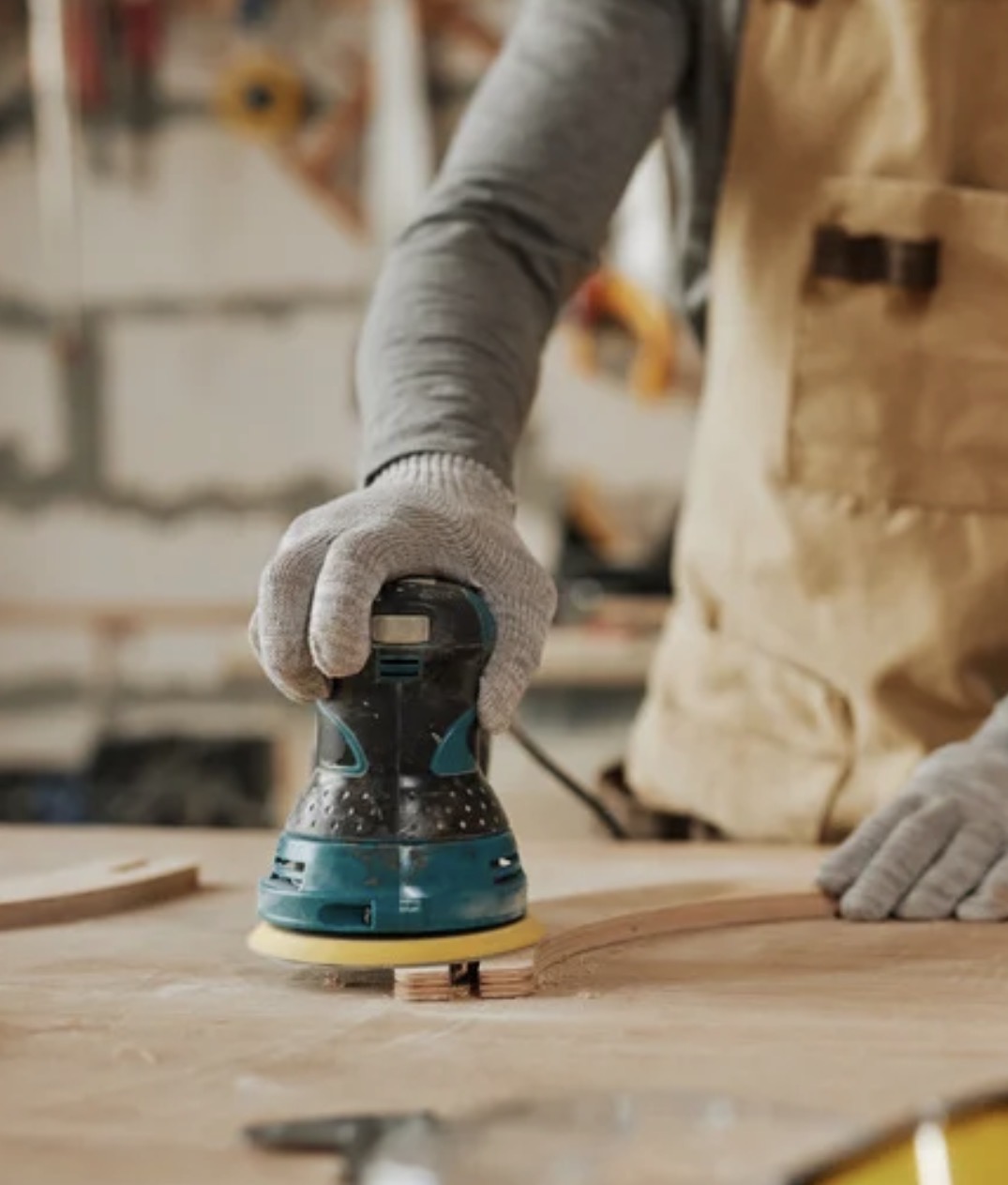 Person using a handheld electric sander on a wooden surface, wearing gloves and a beige apron, in a workshop setting.