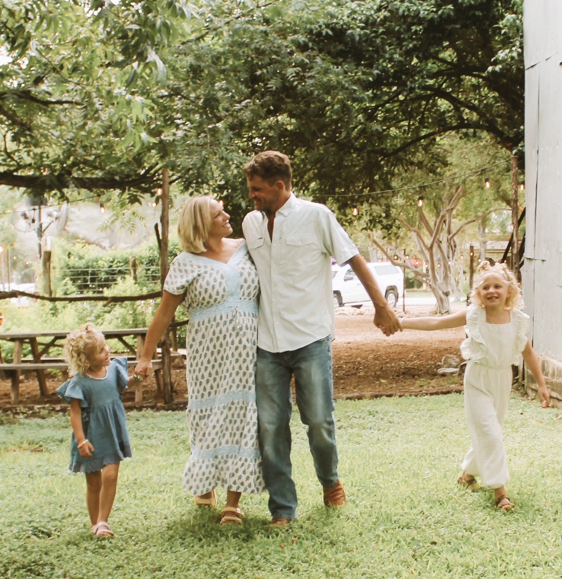 Family of four holding hands and playing outdoors in a park with trees and grass.