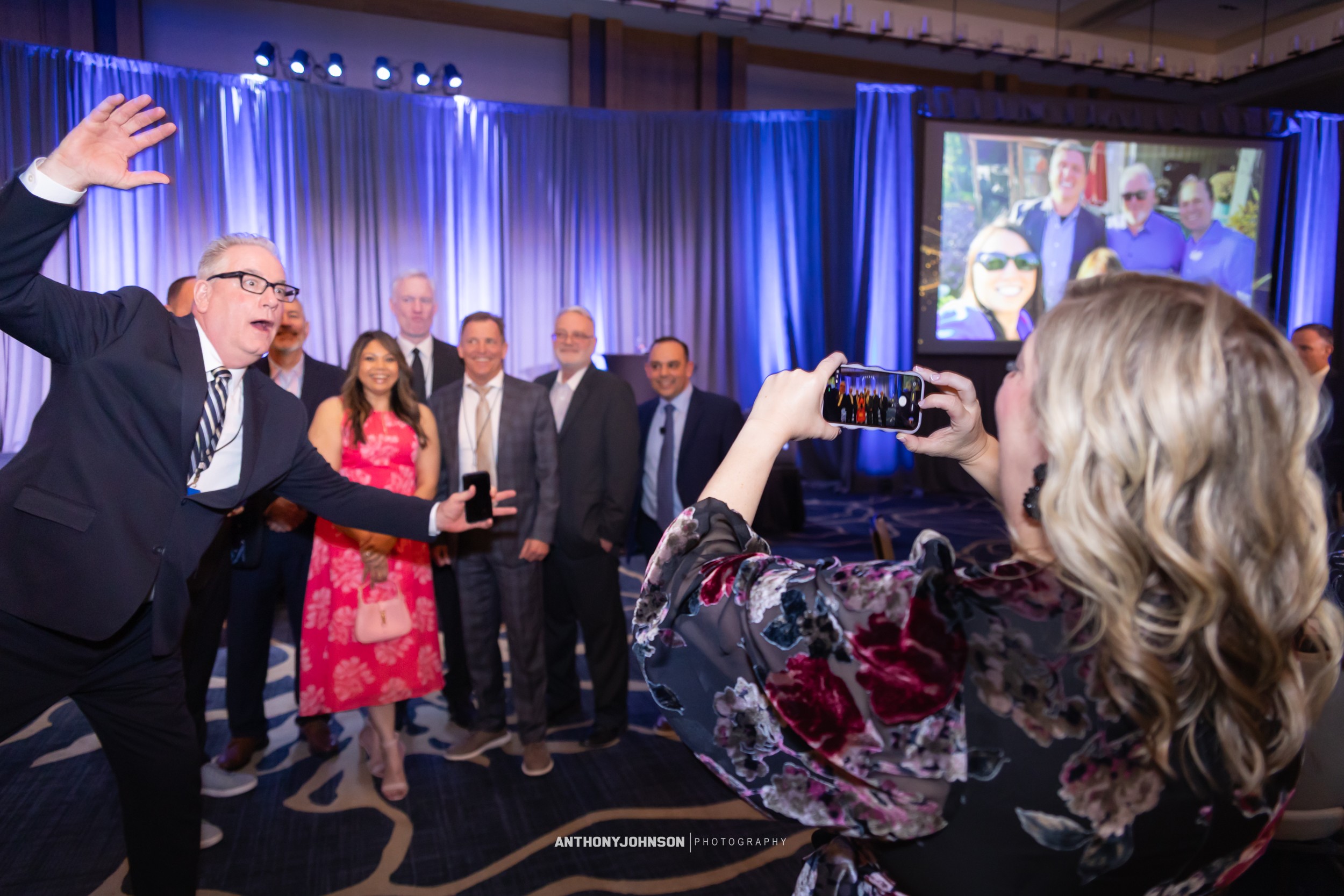 Group of people posing for a photo at an event, with a woman taking a picture using her phone, and a large screen in the background.