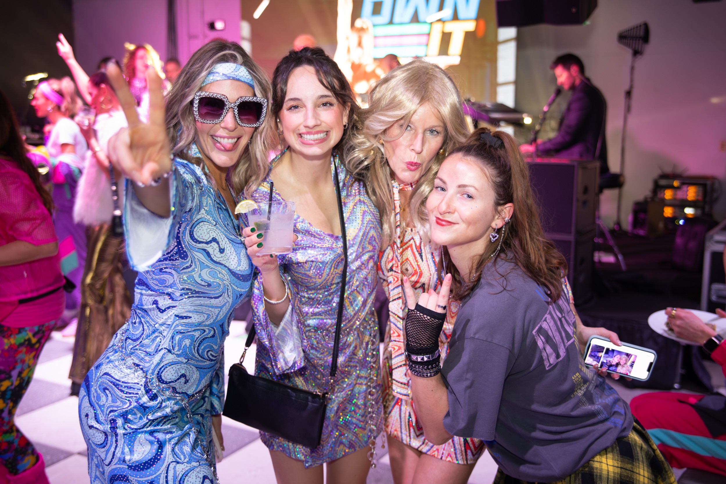 Four women smiling and posing together at a lively party or event, with colorful lights and people in the background.