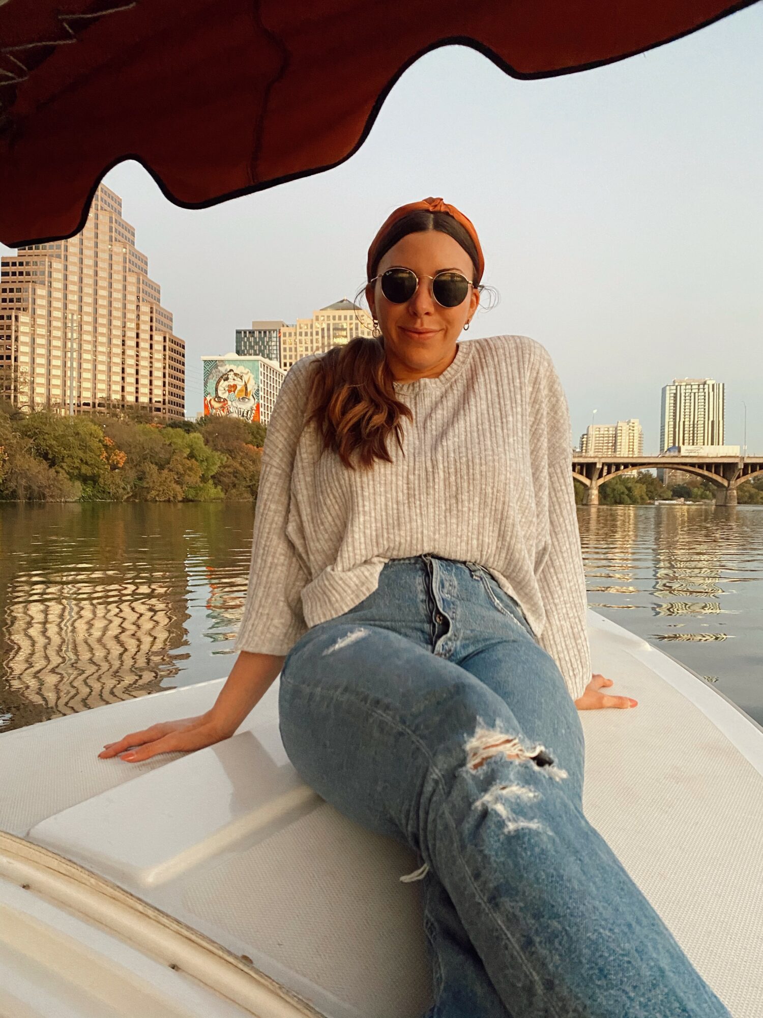 Woman wearing sunglasses and a sweater, sitting on a boat with city skyline and river in background.