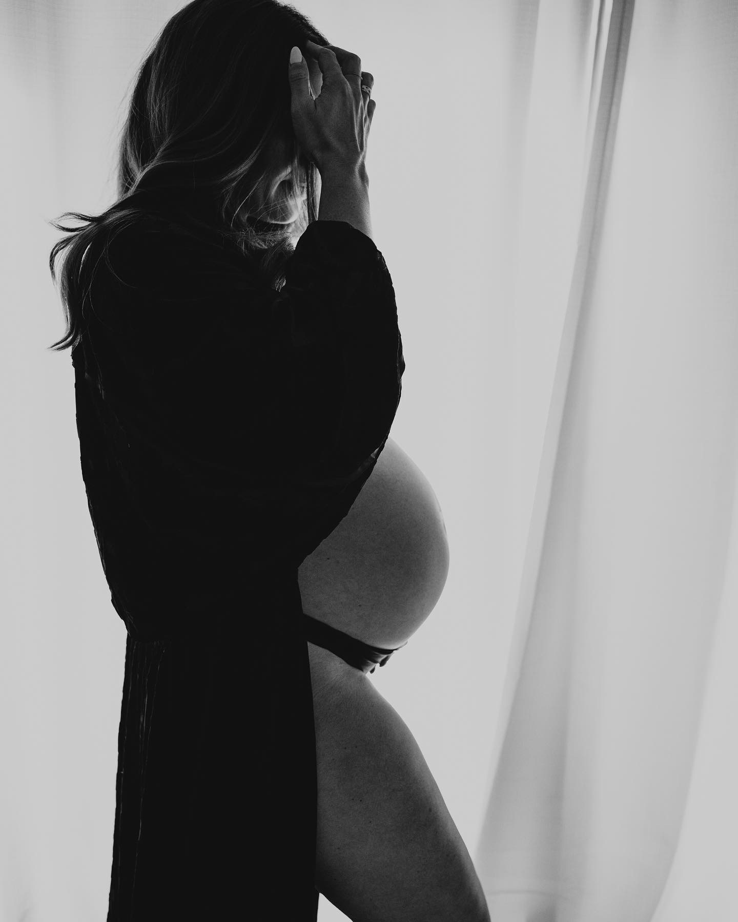 Pregnant woman standing near a window, holding her head, in black and white, side view.