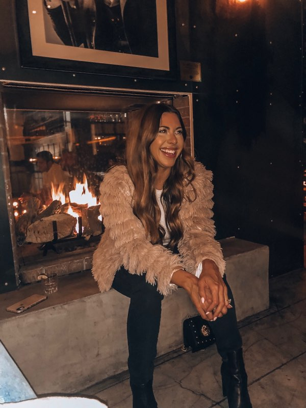 Woman with long hair smiling, sitting by a fireplace in a dimly lit room, wearing a beige fuzzy jacket and black pants.