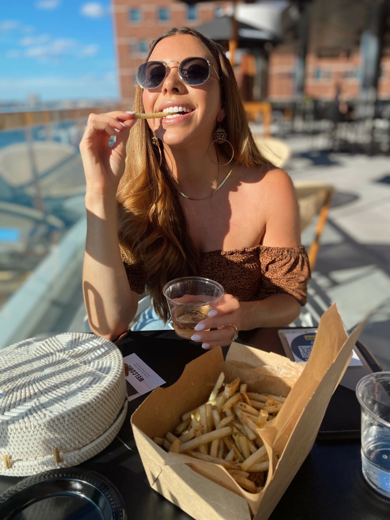 Woman with sunglasses smiling, holding a fry and a drink, outdoor dining with food and drinks on table.