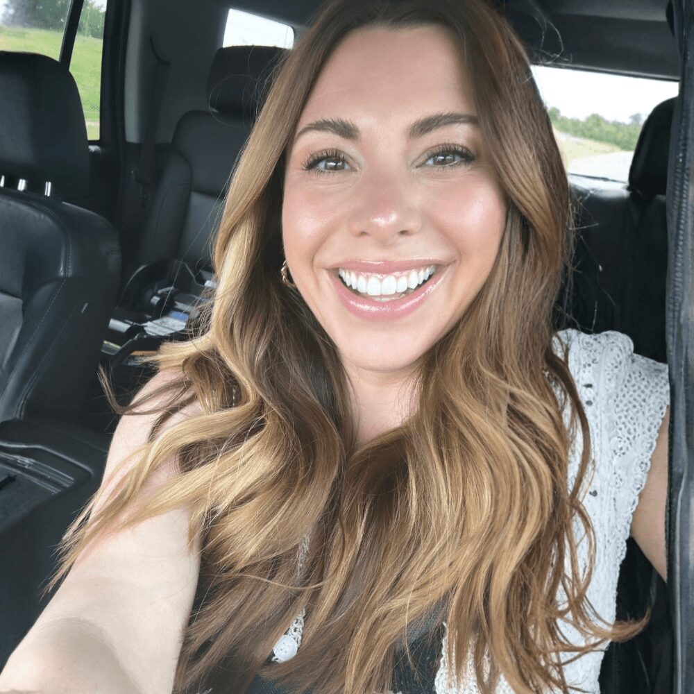 Smiling woman with long wavy hair in car, wearing a white top, bright expression, natural light, background of car interior.
