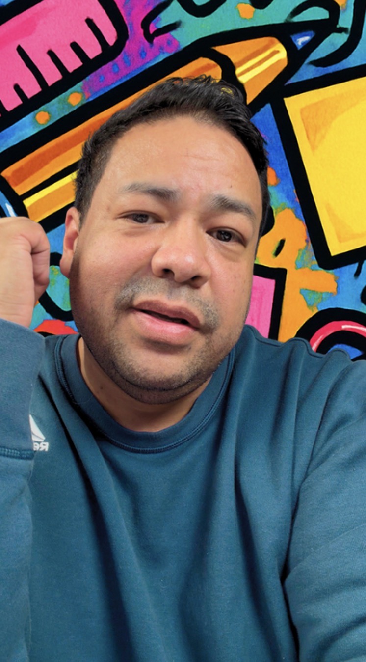 Man with short dark hair and a blue shirt taking a selfie in front of a colorful graffiti wall.